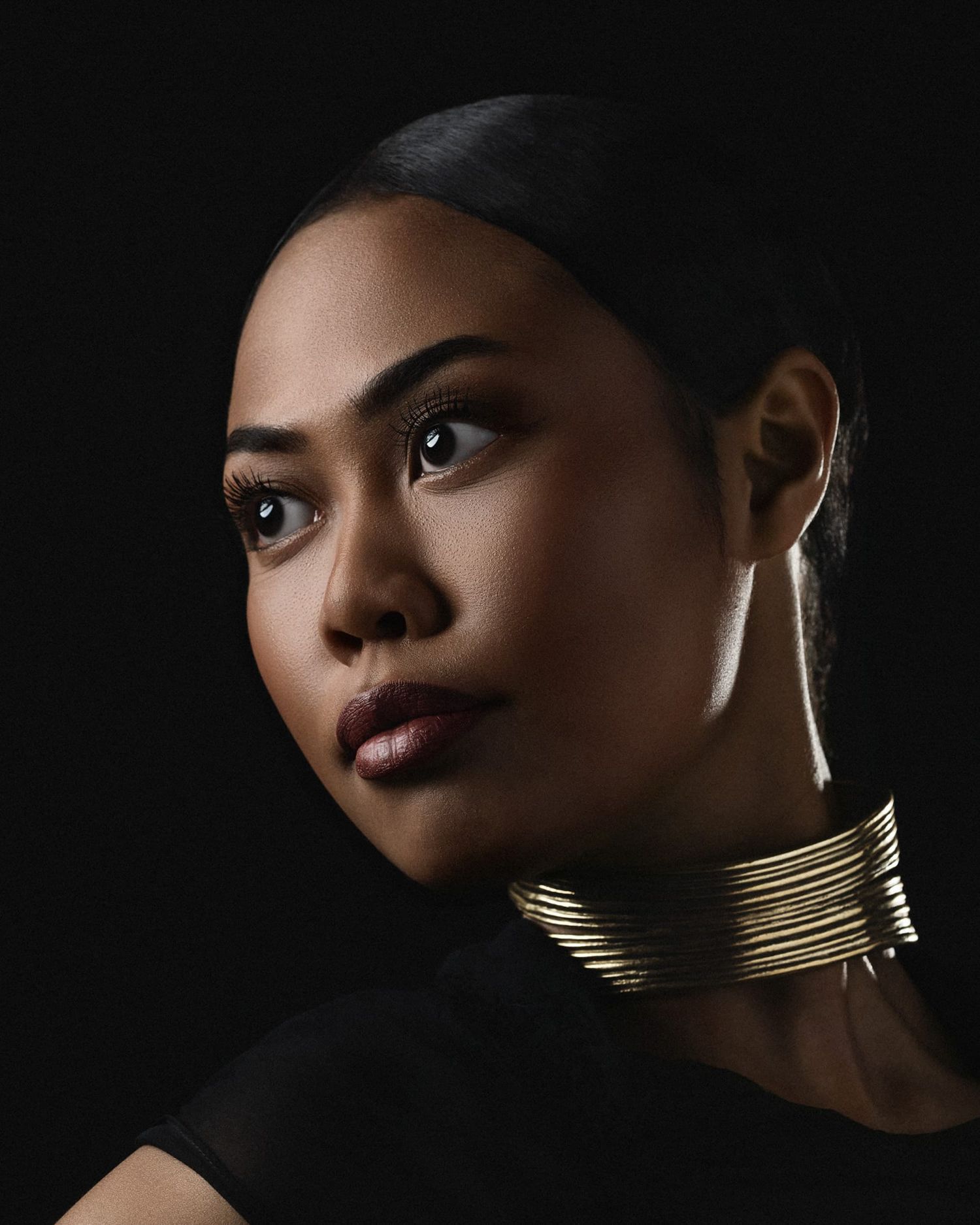Woman wearing a gold coil choker necklace against a dark background in dramatic studio lighting.