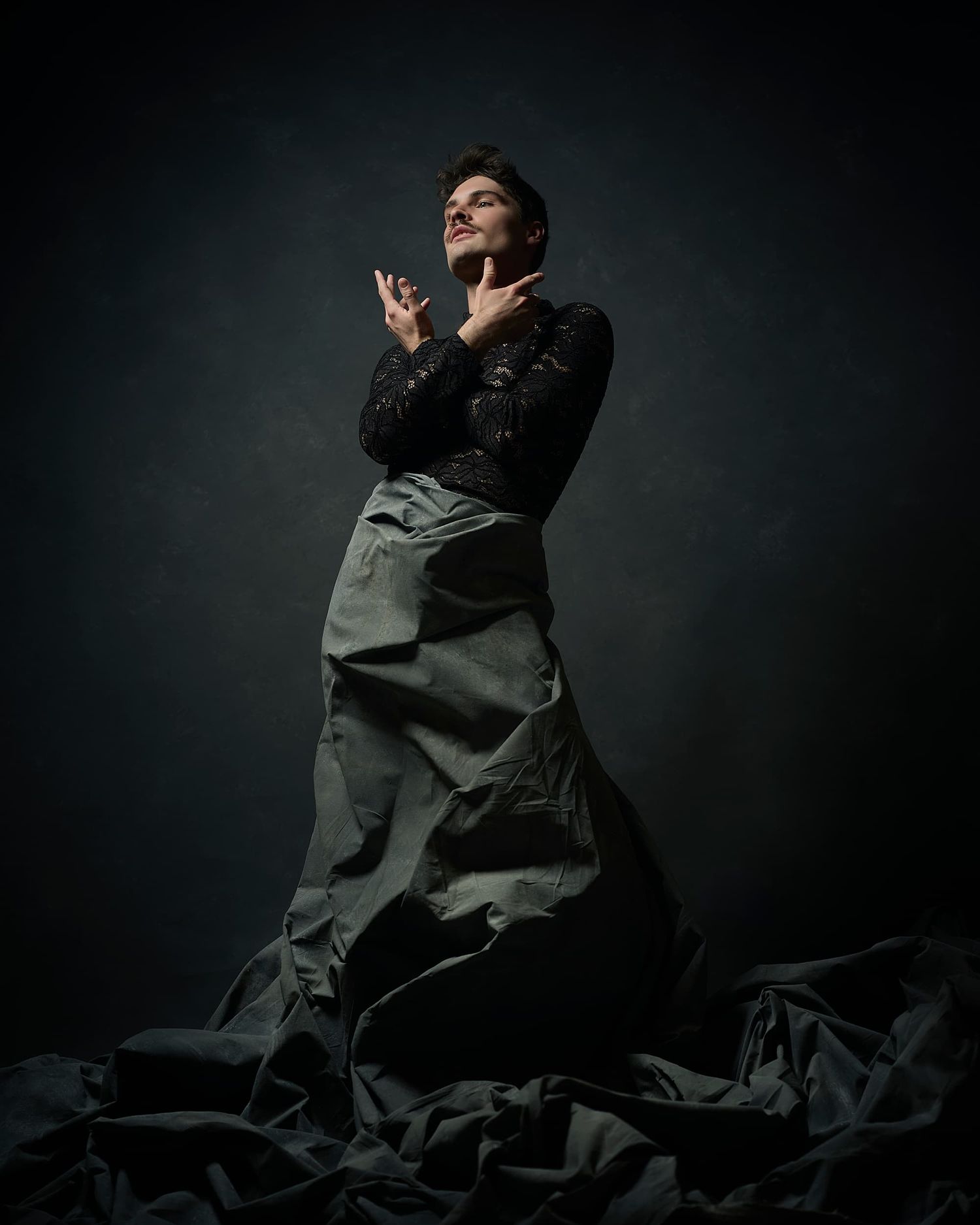 Person in a black top and gray skirt poses dramatically against a dark moody studio backdrop.