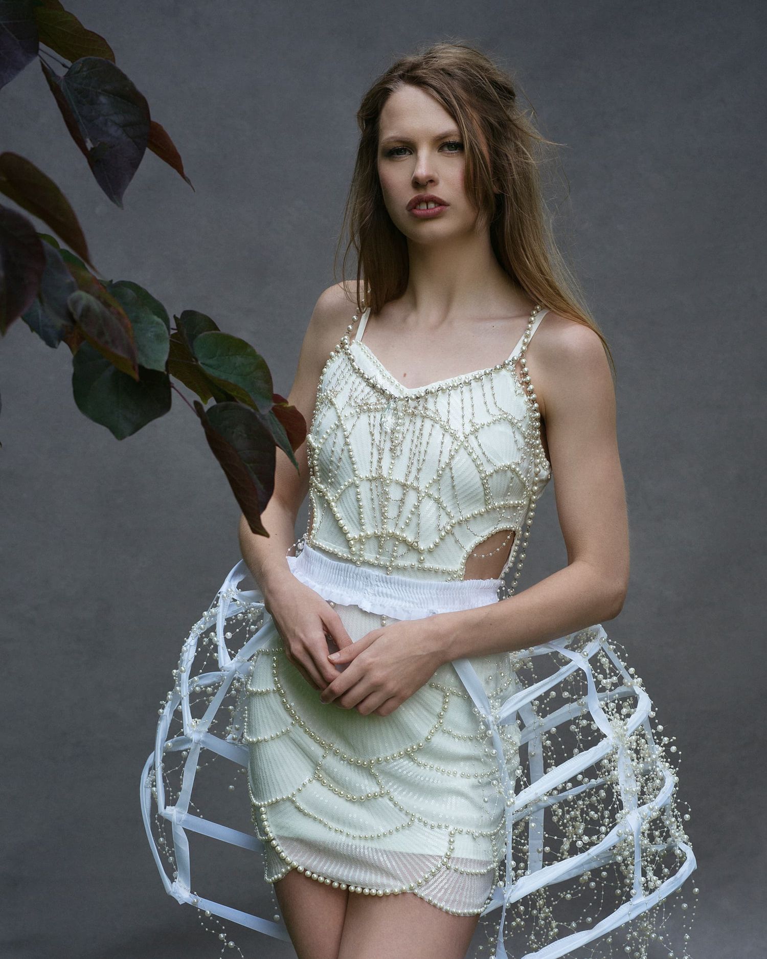 Model in a pearl-embellished white mini dress with hoop skirt beside a plant.