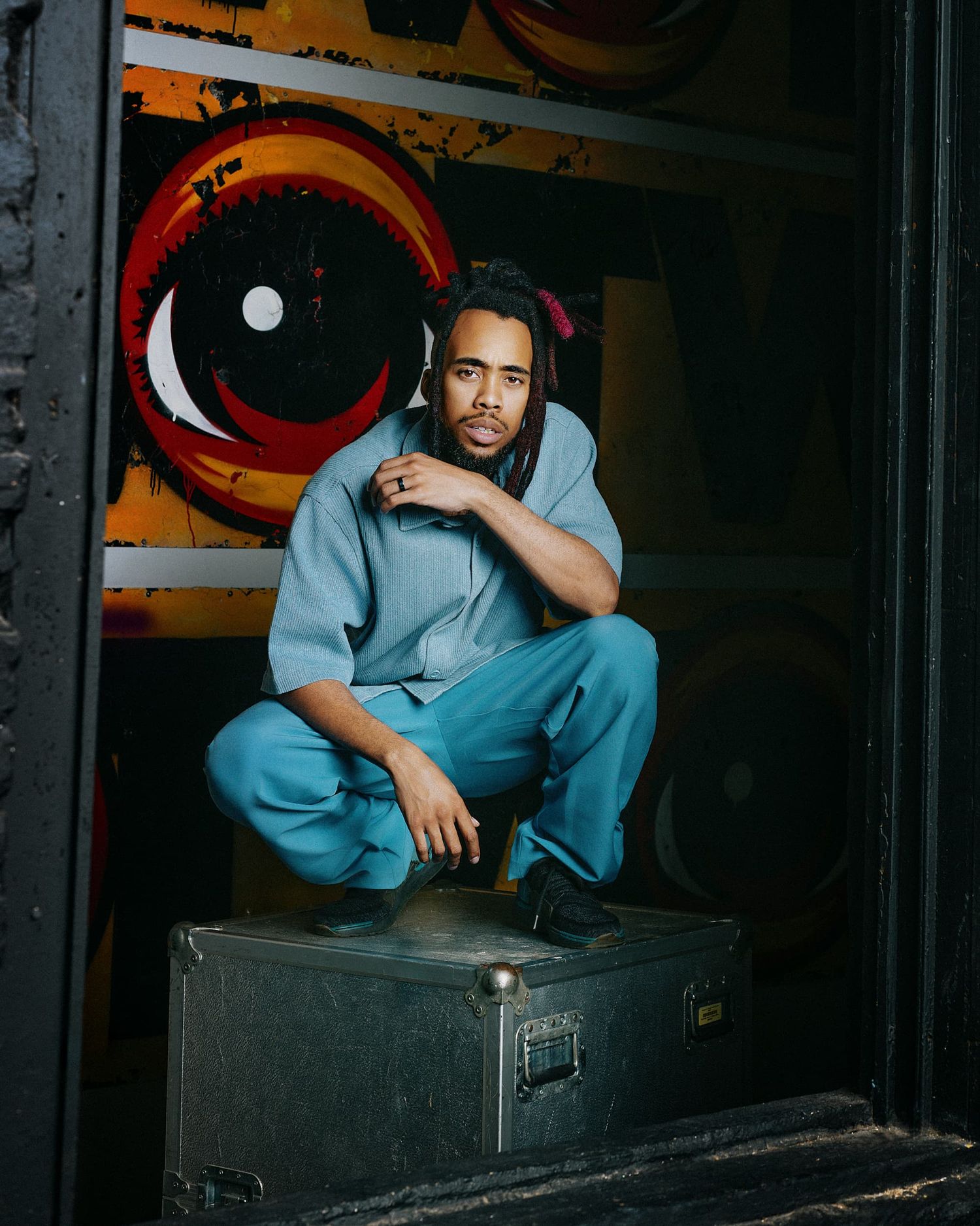 Young man in a blue hoodie and pants crouching on a road case in front of colorful graffiti artwork.