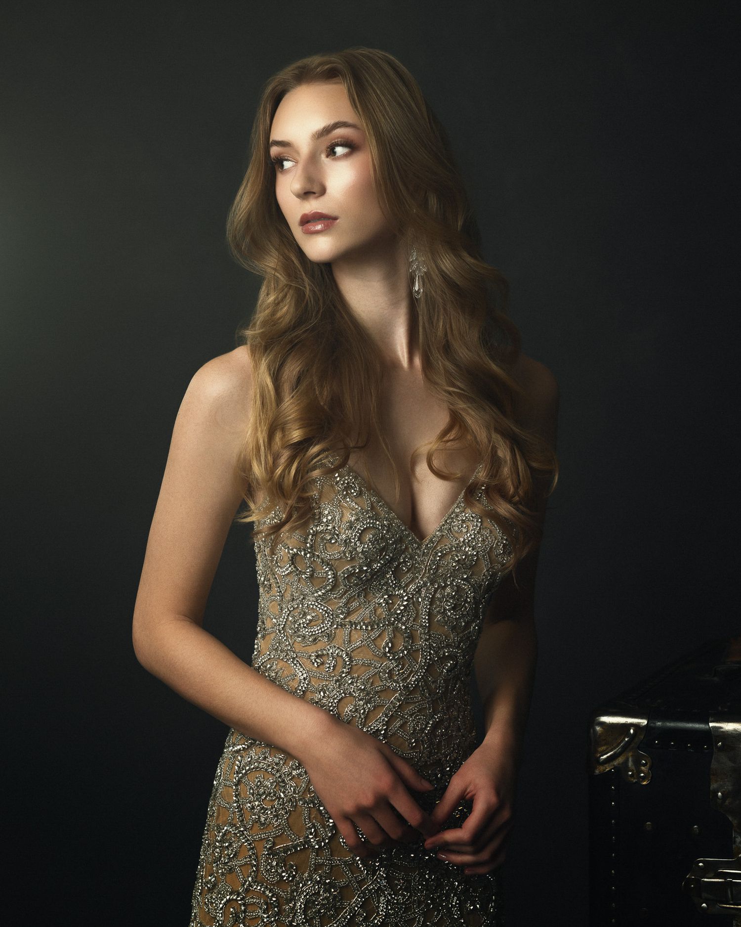 Young woman in ornate beaded gown with long wavy hair poses in dark studio setting.