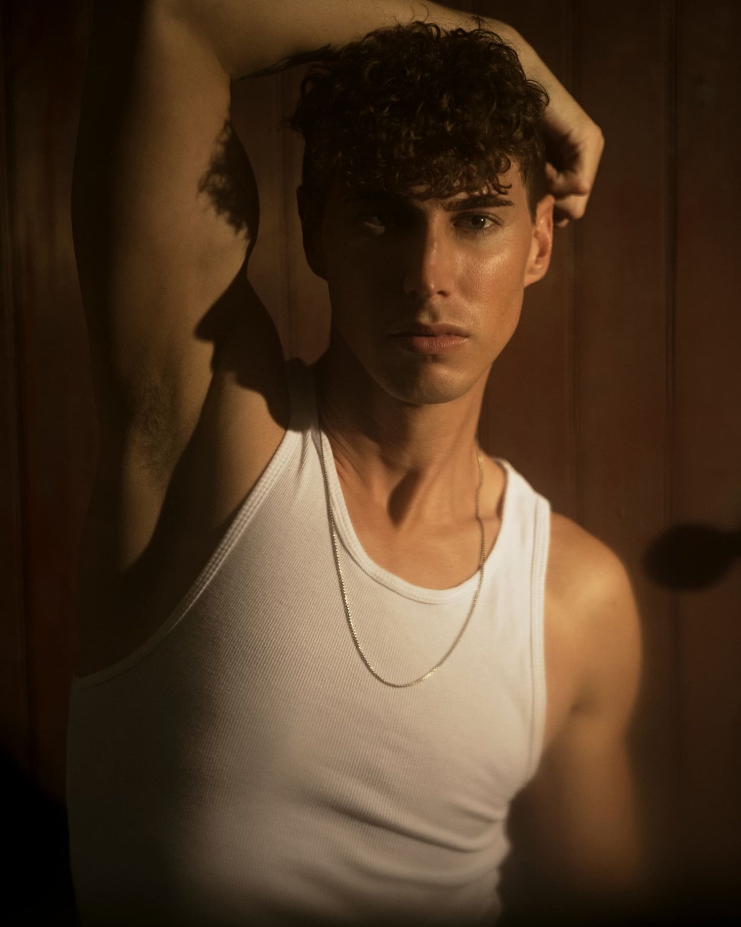 Young man with curly hair in a white tank top and chain necklace poses in warm golden light.