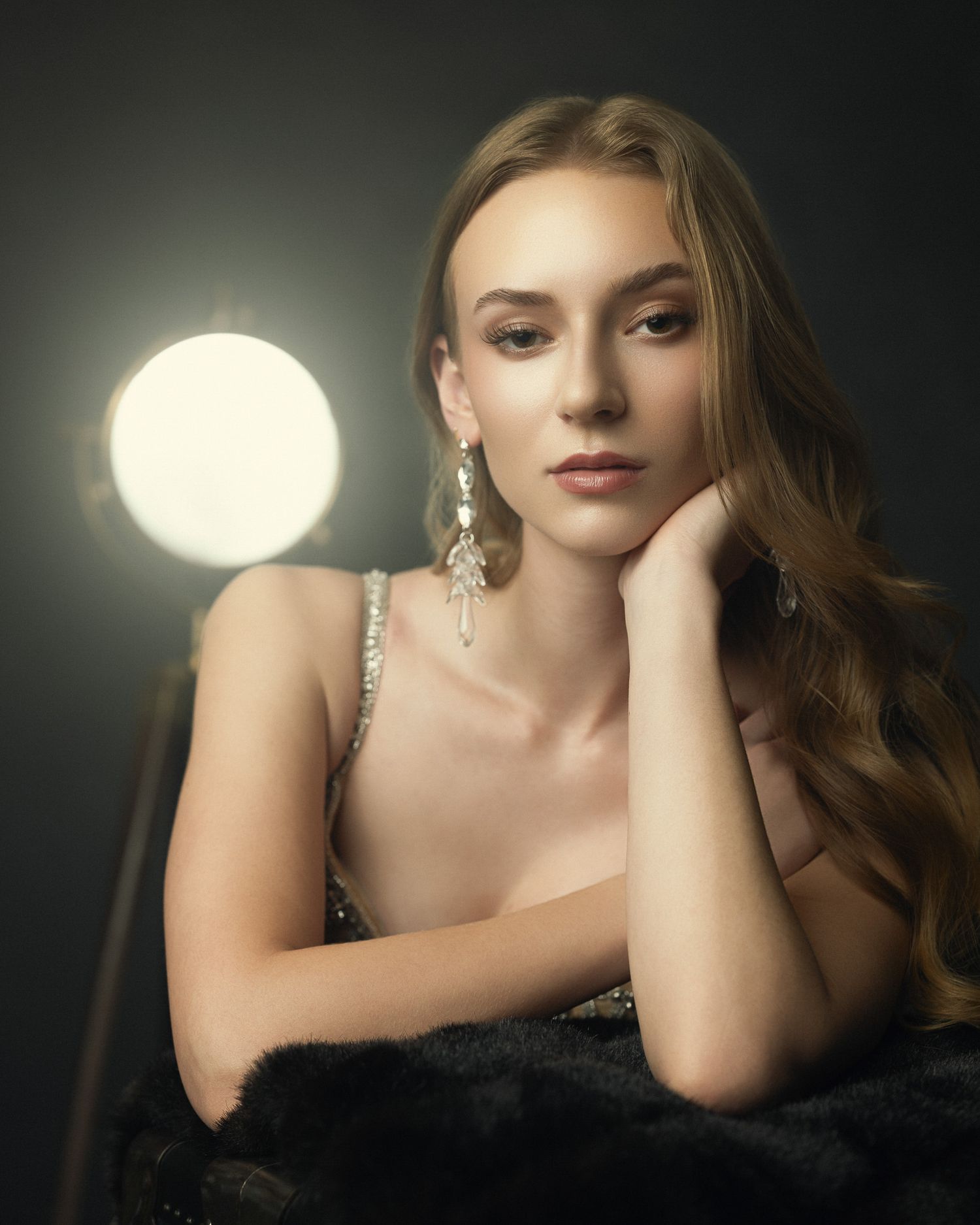 Elegant woman in sequined dress with silver earrings rests chin on hand near studio light.