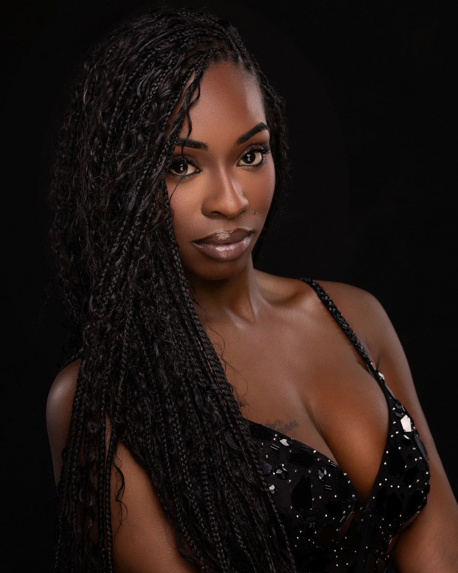 Woman with long box braids wearing a black sequined top poses against a dark studio background.