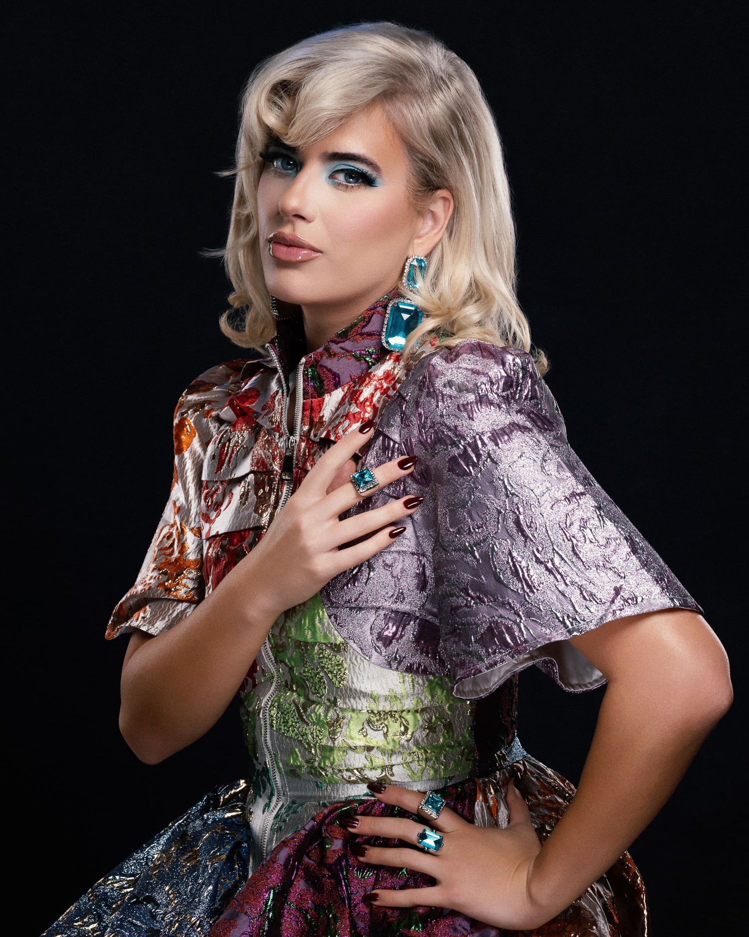 Model in a colorful metallic patchwork outfit with teal earrings and dark nails.