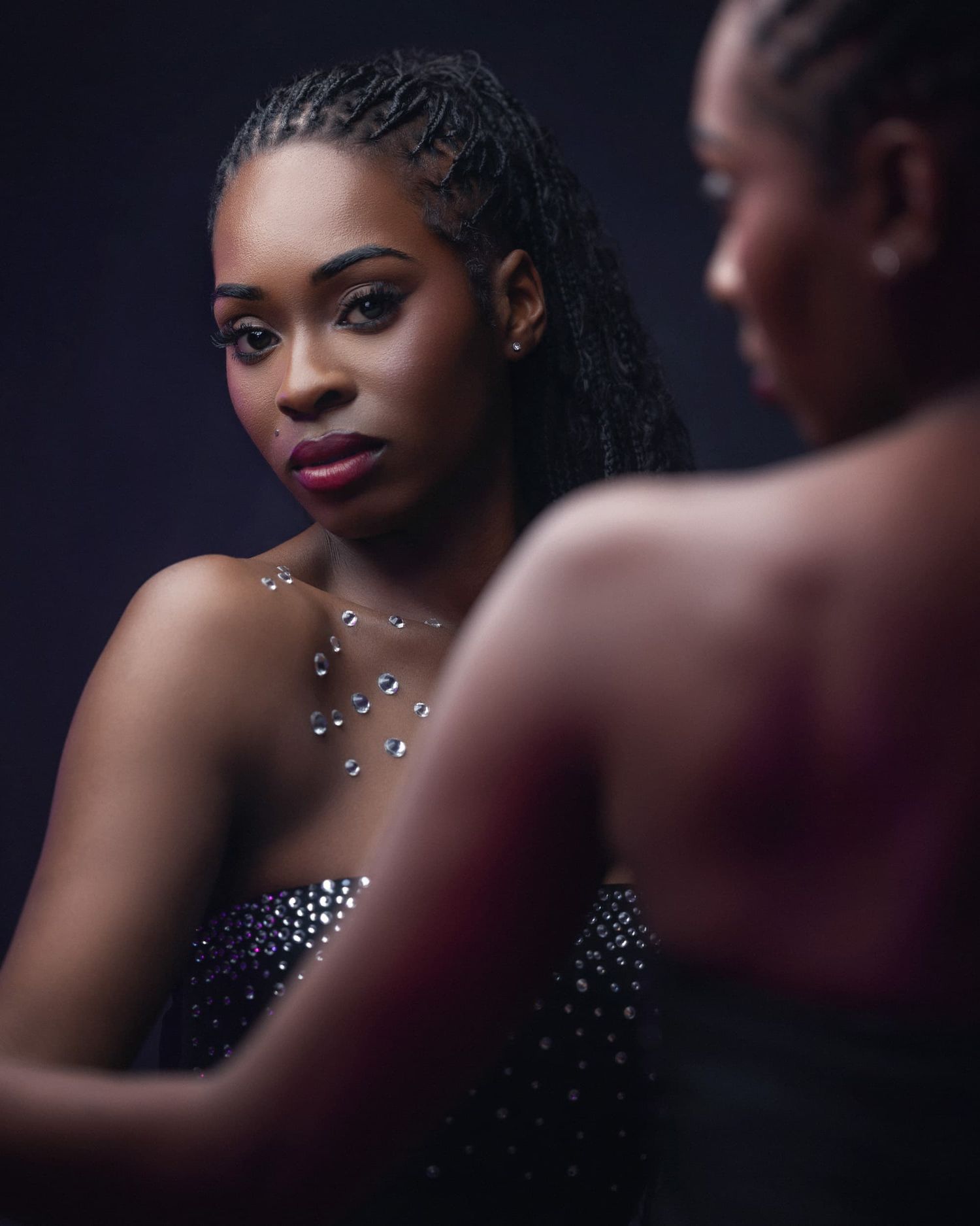 Young woman with braided hair wearing a sparkling necklace and dark outfit reflected in a mirror.