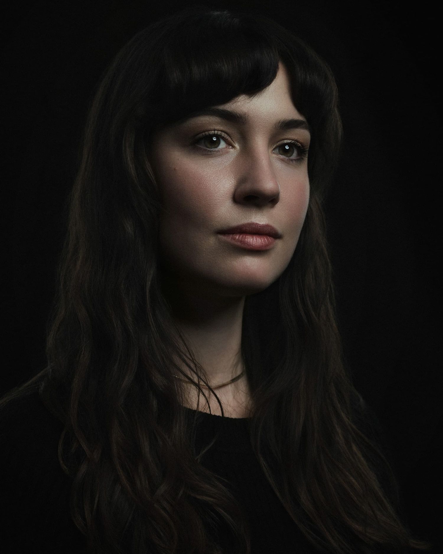 Dark portrait of a young woman with long dark hair and bangs against a black background.