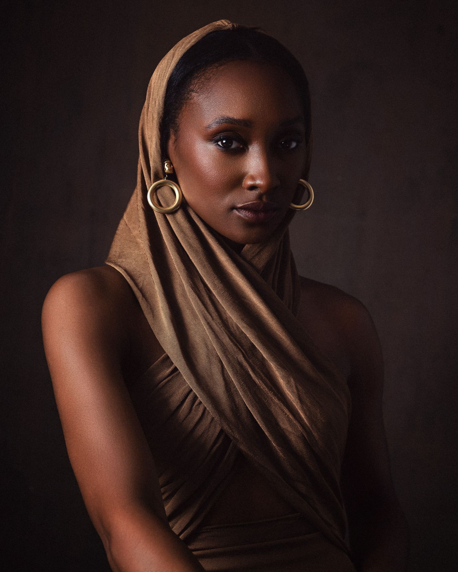 A Black woman draped in a tan headwrap and matching outfit wears gold hoop earrings against a dark background.