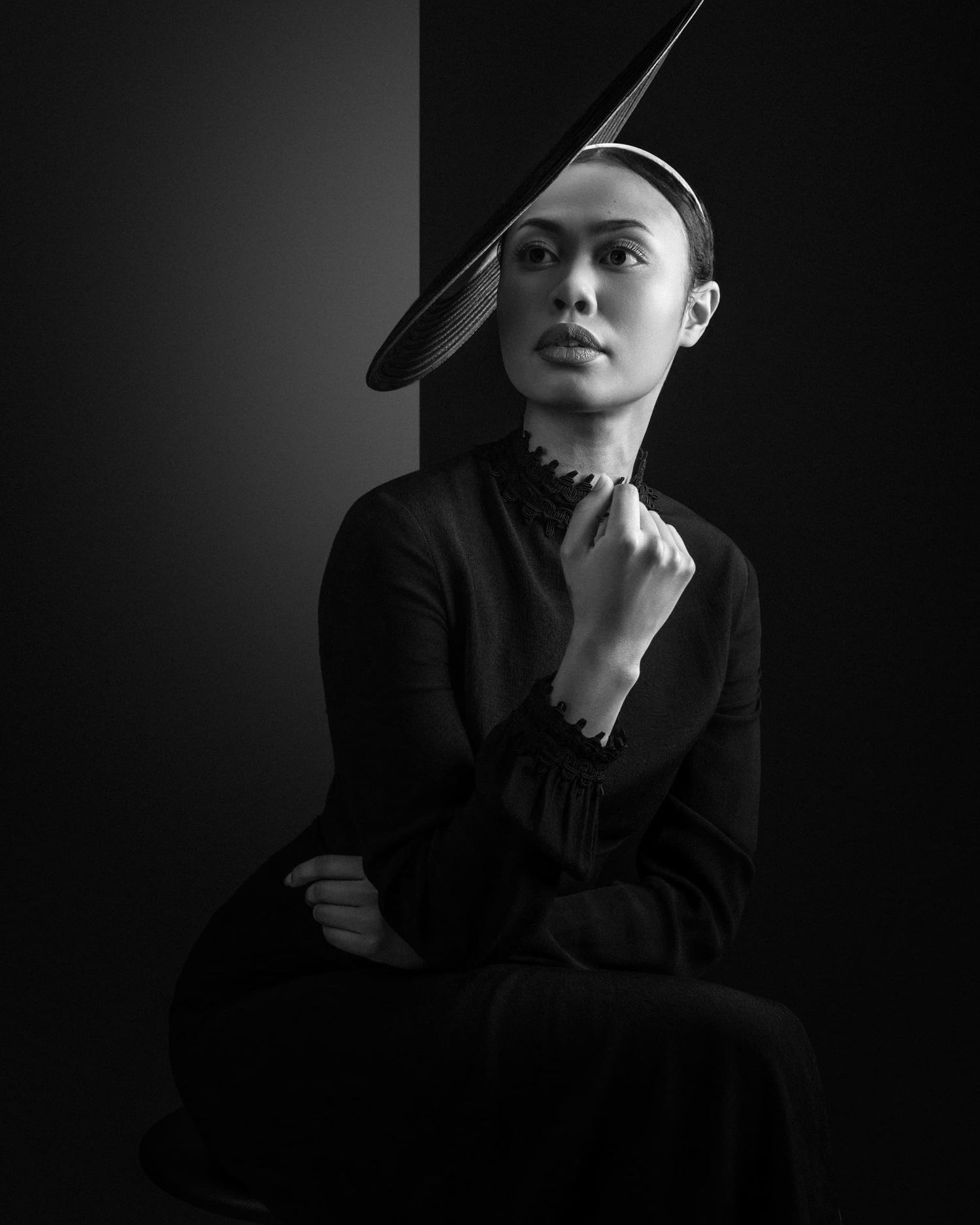 Black and white portrait of a woman in black wearing an avant-garde angular hat in a studio.