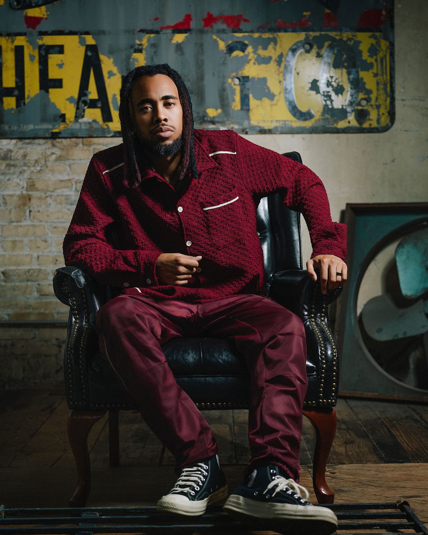 Man in red outfit sits confidently in a leather chair against a rustic brick wall background.