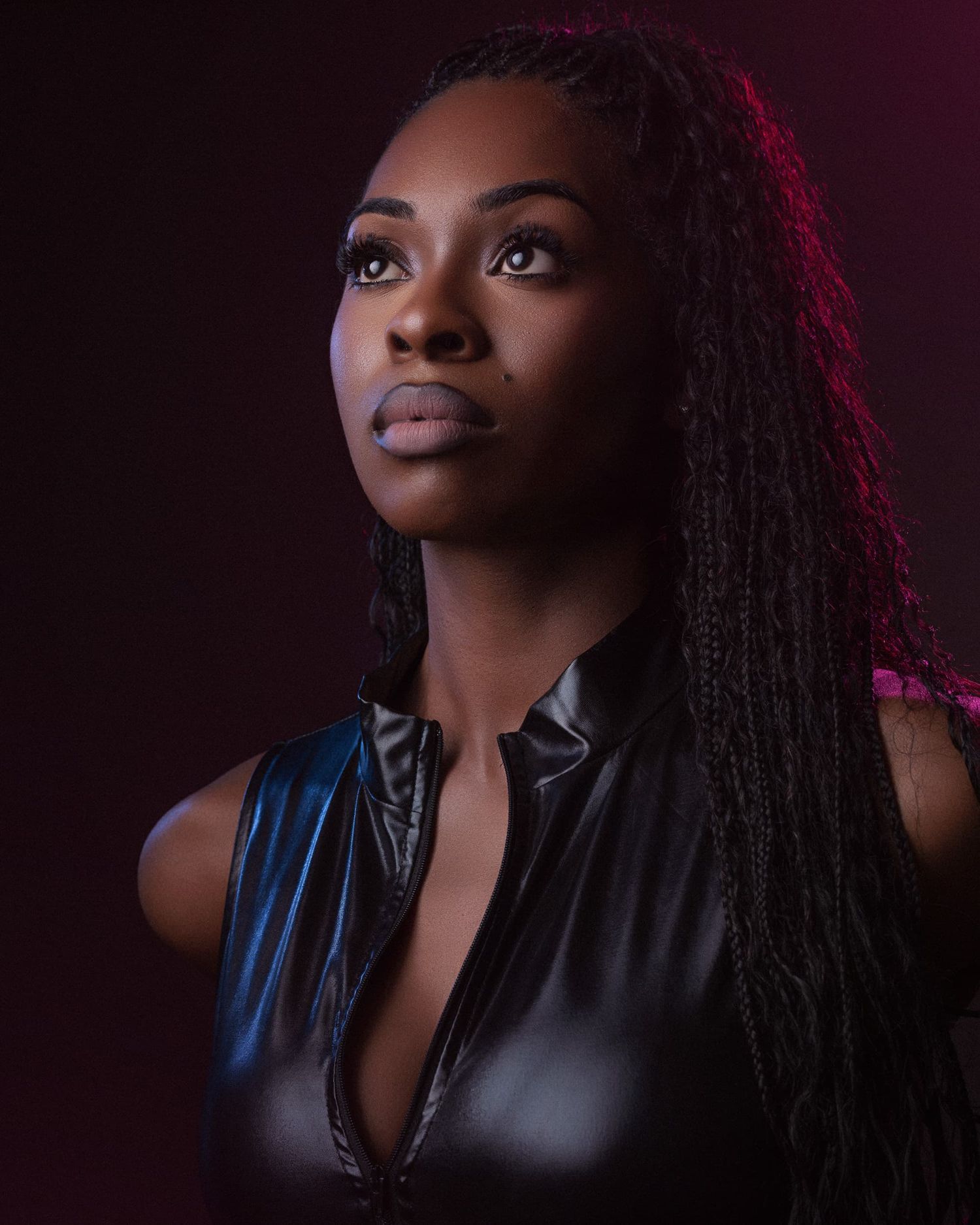 Woman with long braided hair in a black leather top poses under dramatic purple and red lighting.
