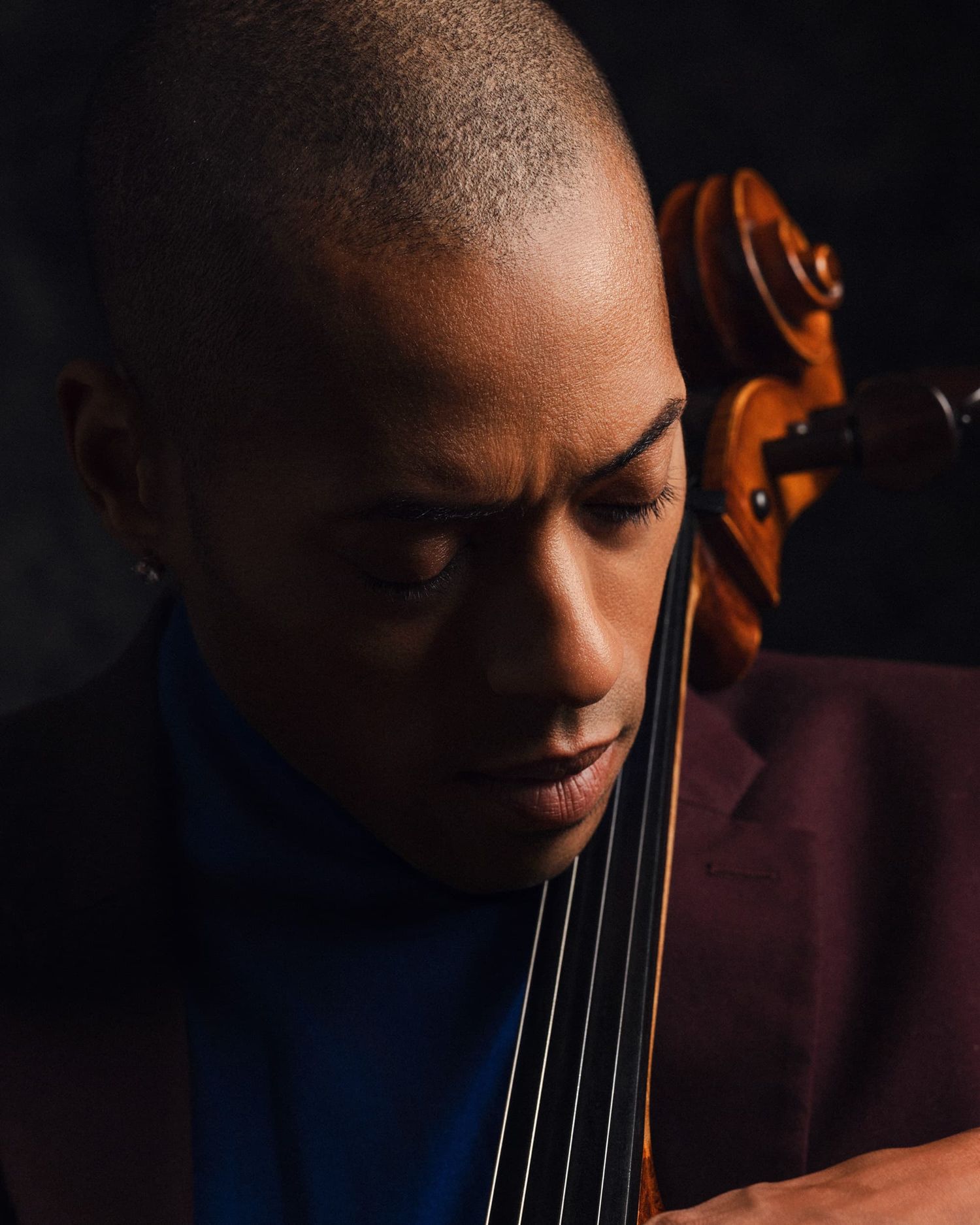 Close-up of a young man with short hair looking down while playing a cello in warm studio lighting.