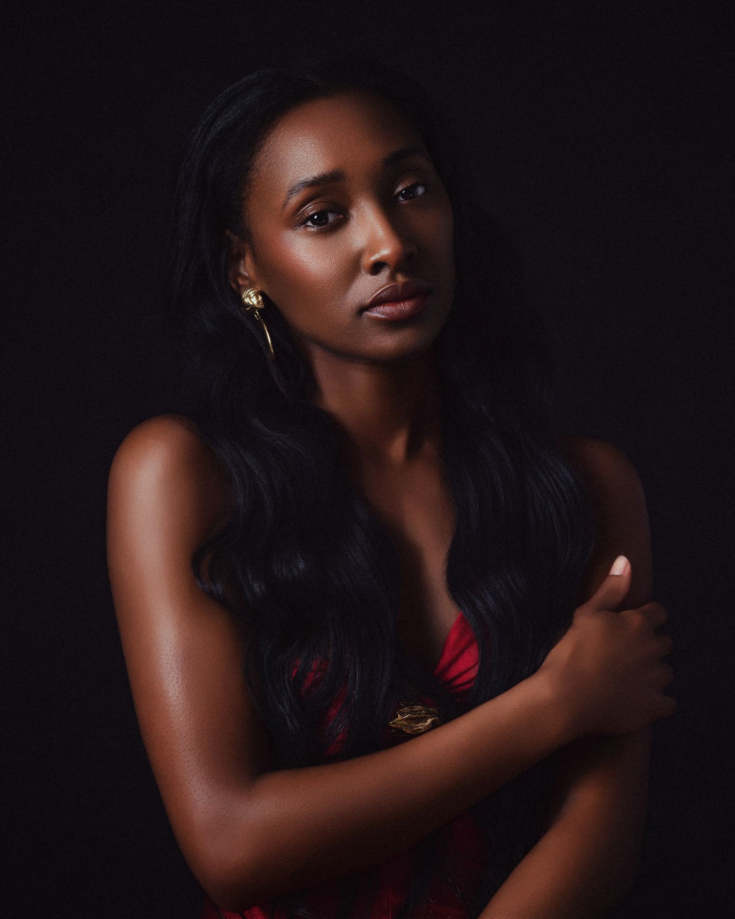 A Black woman in a red floral dress with gold earrings crosses her arms against a dark background.