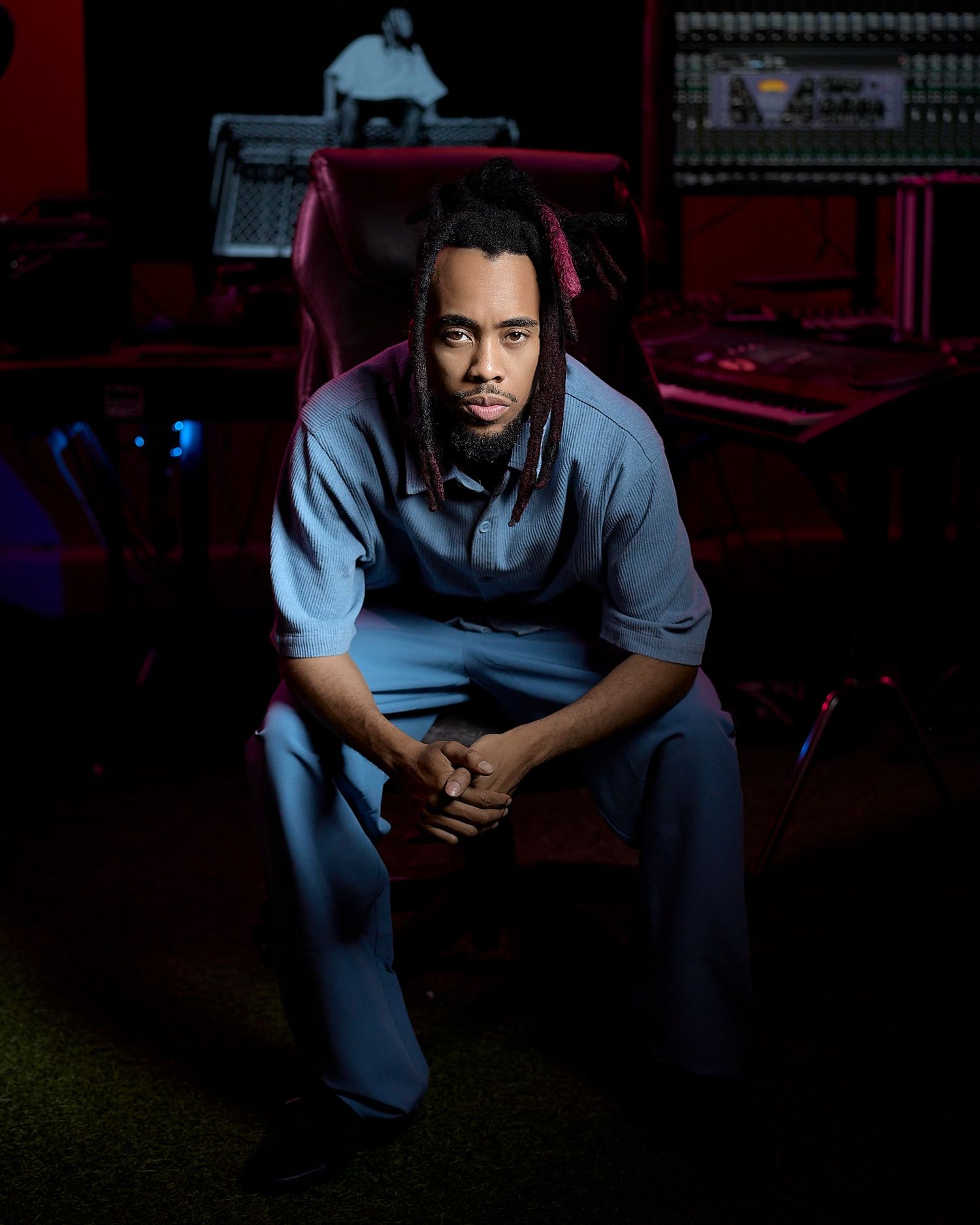 Man with dreadlocks sits in a recording studio chair wearing a blue outfit, surrounded by music equipment.