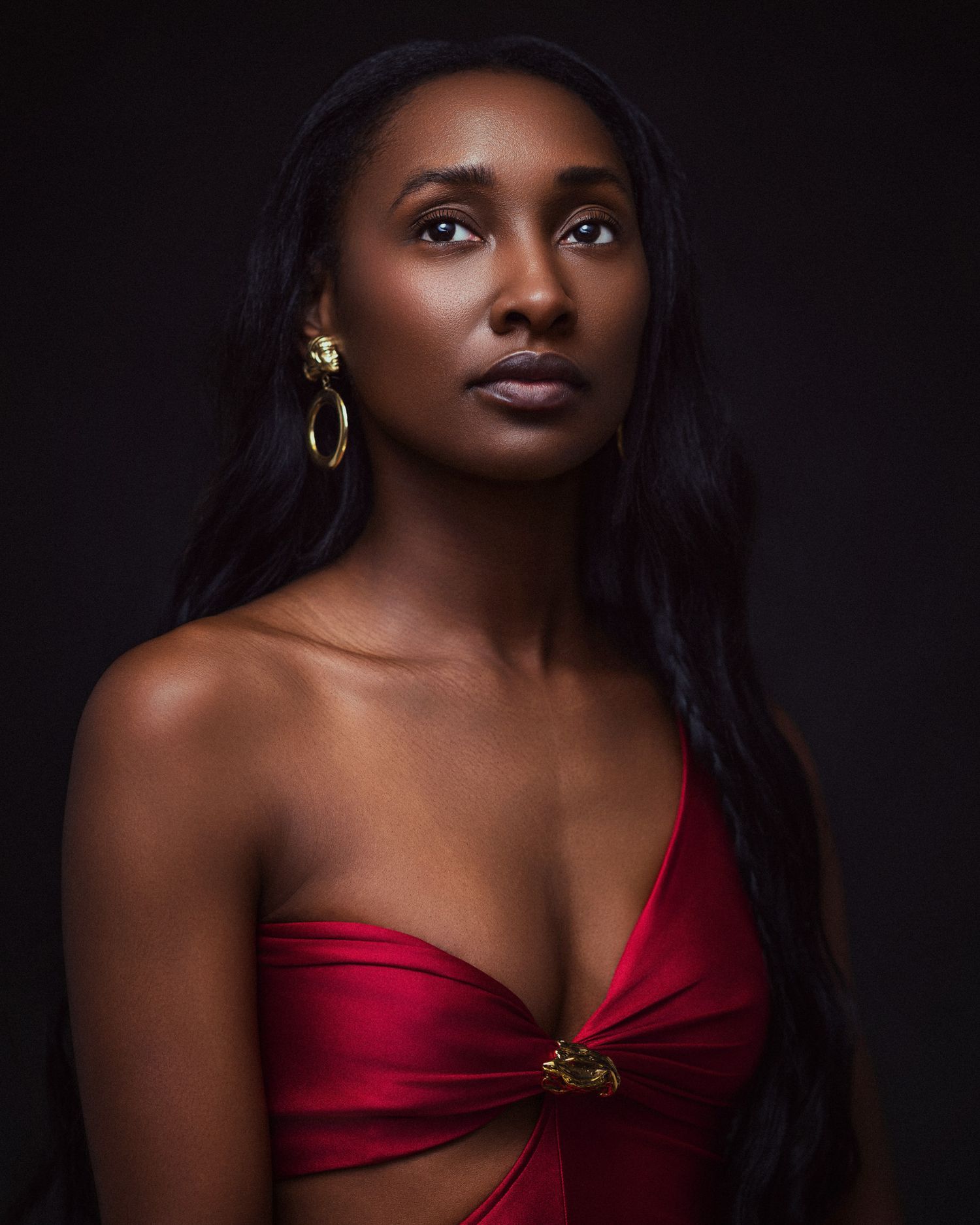 A Black woman in a red strapless top with gold hoop earrings poses against a dark background.