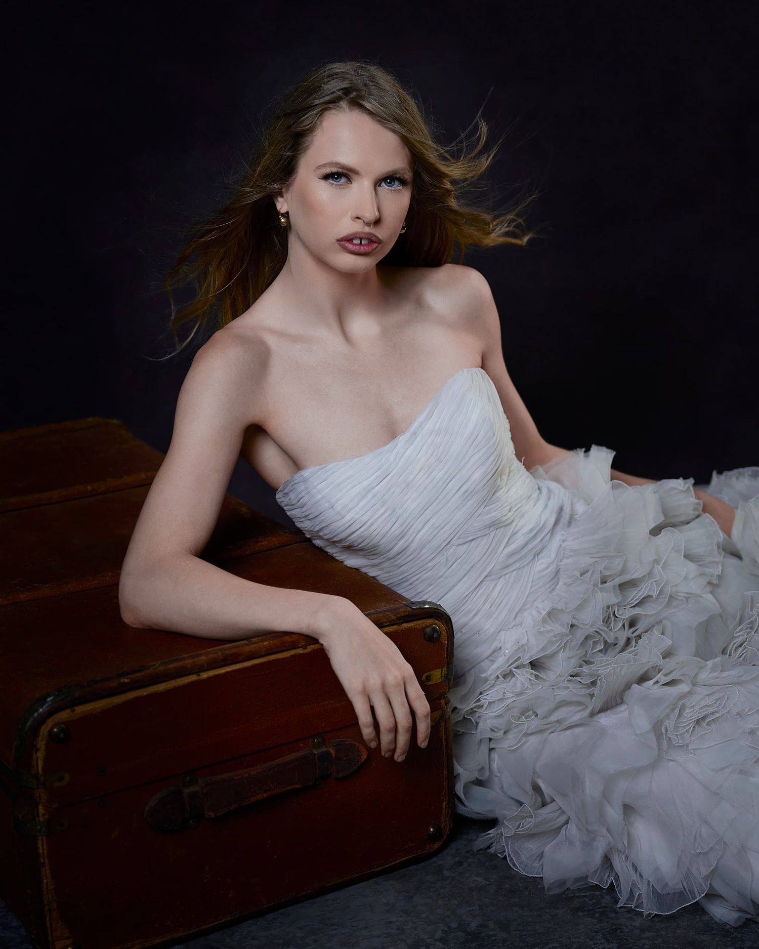 Model in a white strapless ruffled bridal gown leaning on a vintage trunk.