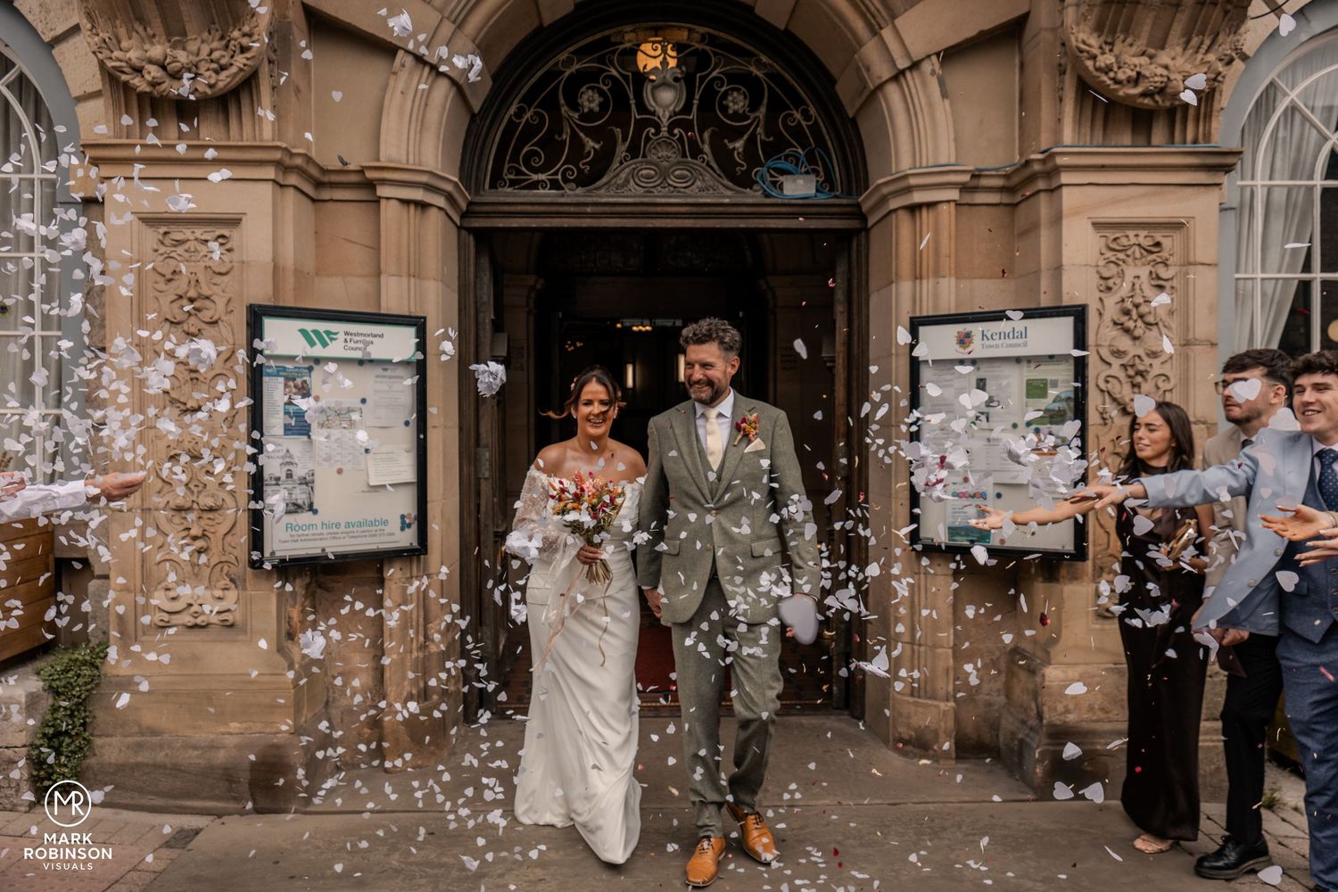 Bride and groom leaving an ornate wedding venue as guests throw confetti around them.
