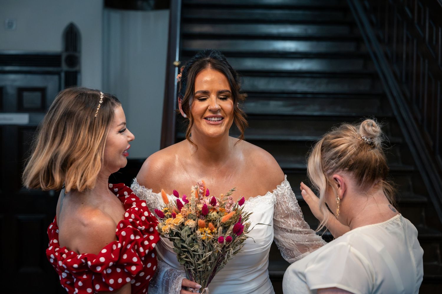 Smiling bride holding a colourful bouquet while two bridesmaids help her prepare beside a staircase.