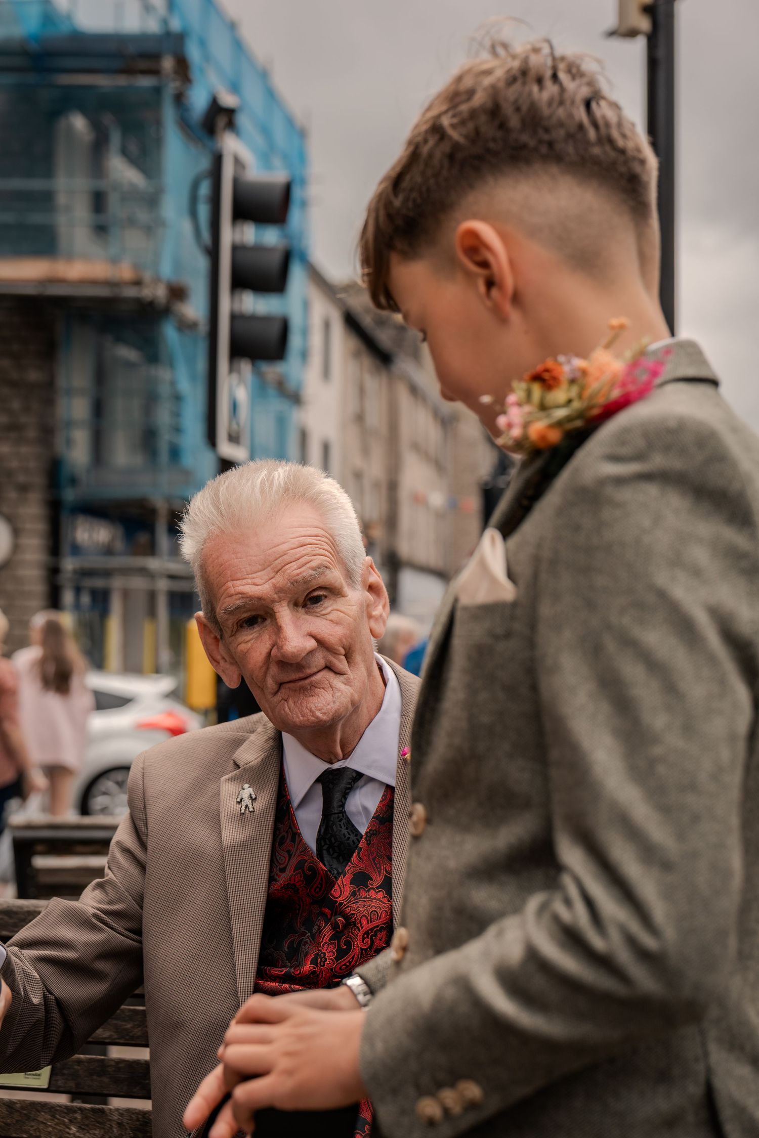 Young wedding guest in a grey suit speaking with an older man outside the venue. Granddad with Grandson