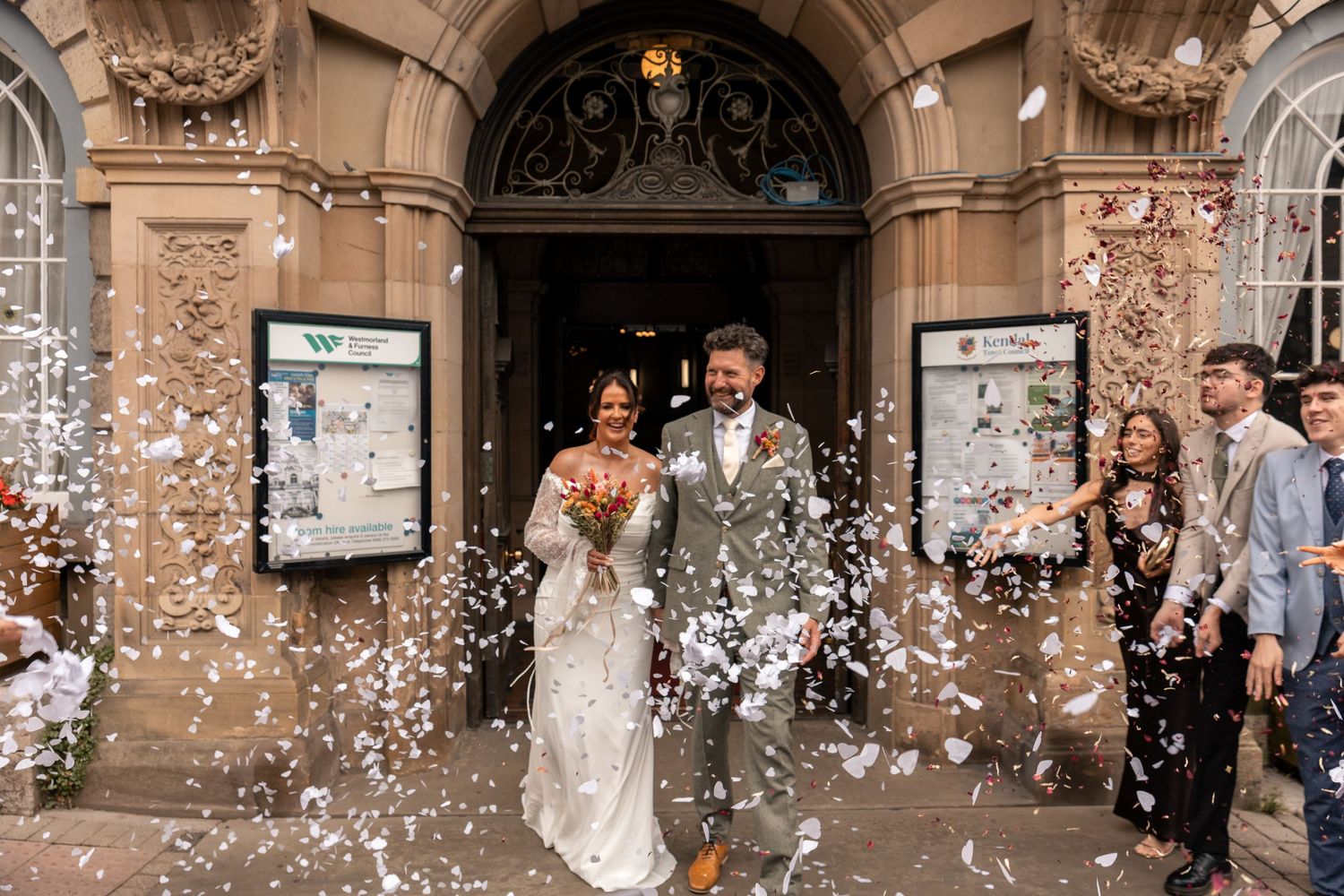Bride and groom leaving an ornate wedding venue as guests throw confetti around them.