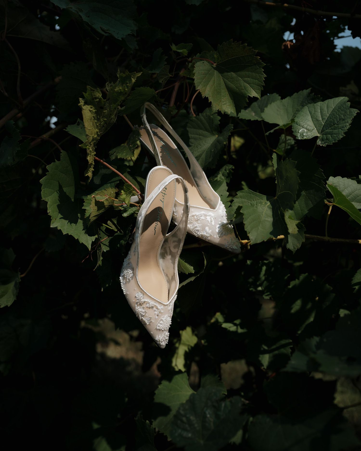 Elegant white lace bridal heels hanging among lush green leaves in natural outdoor setting.