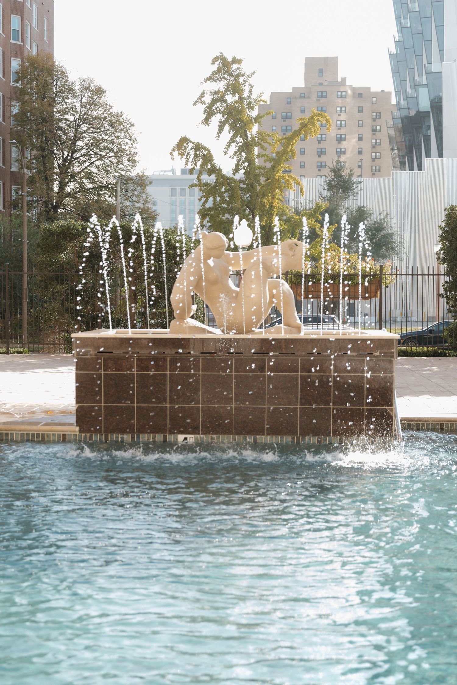Ornate stone fountain sculpture with water jets in urban plaza surrounded by city buildings and trees.
