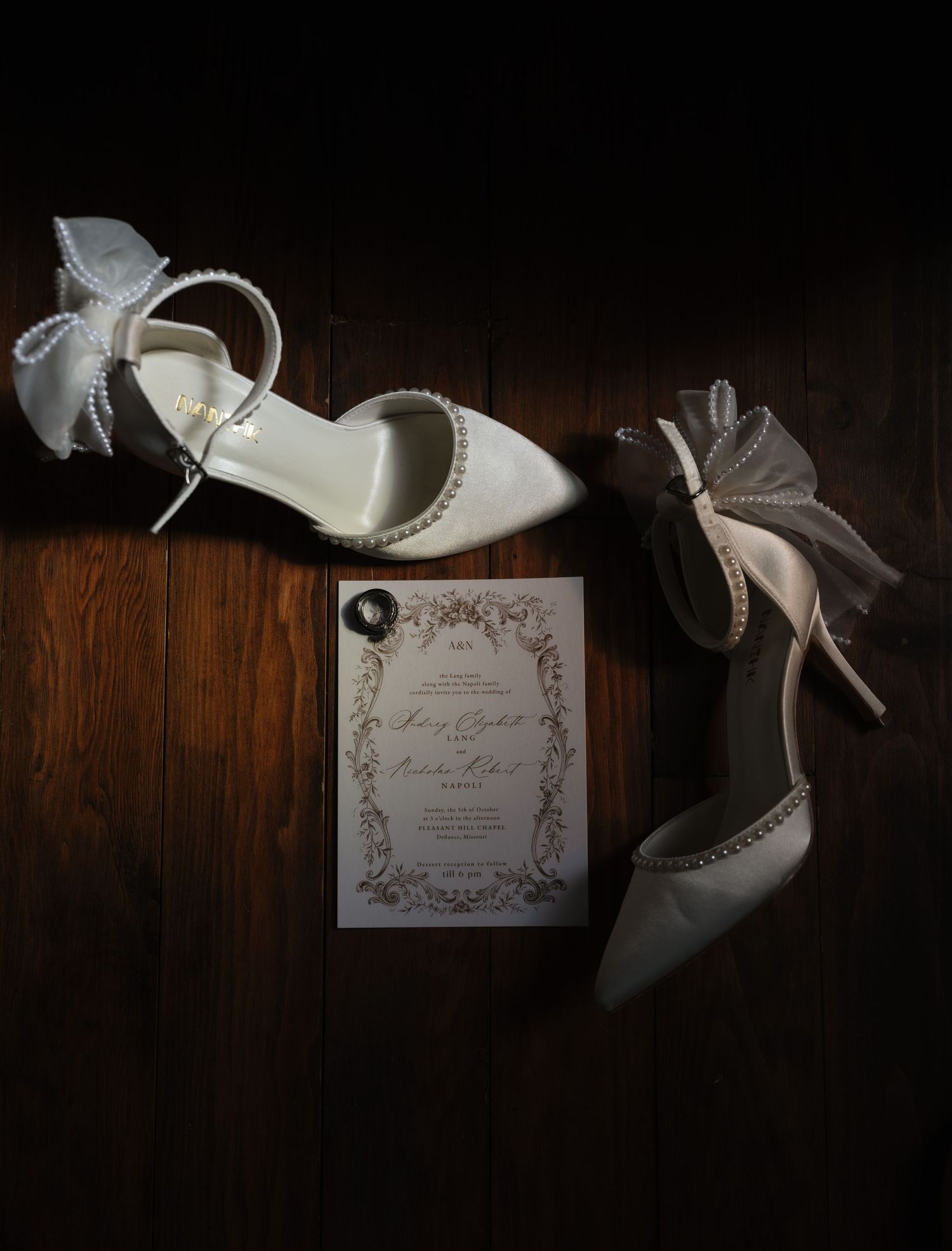 White pearl-embellished bridal heels with bow detail beside elegant wedding invitation on dark wood floor.