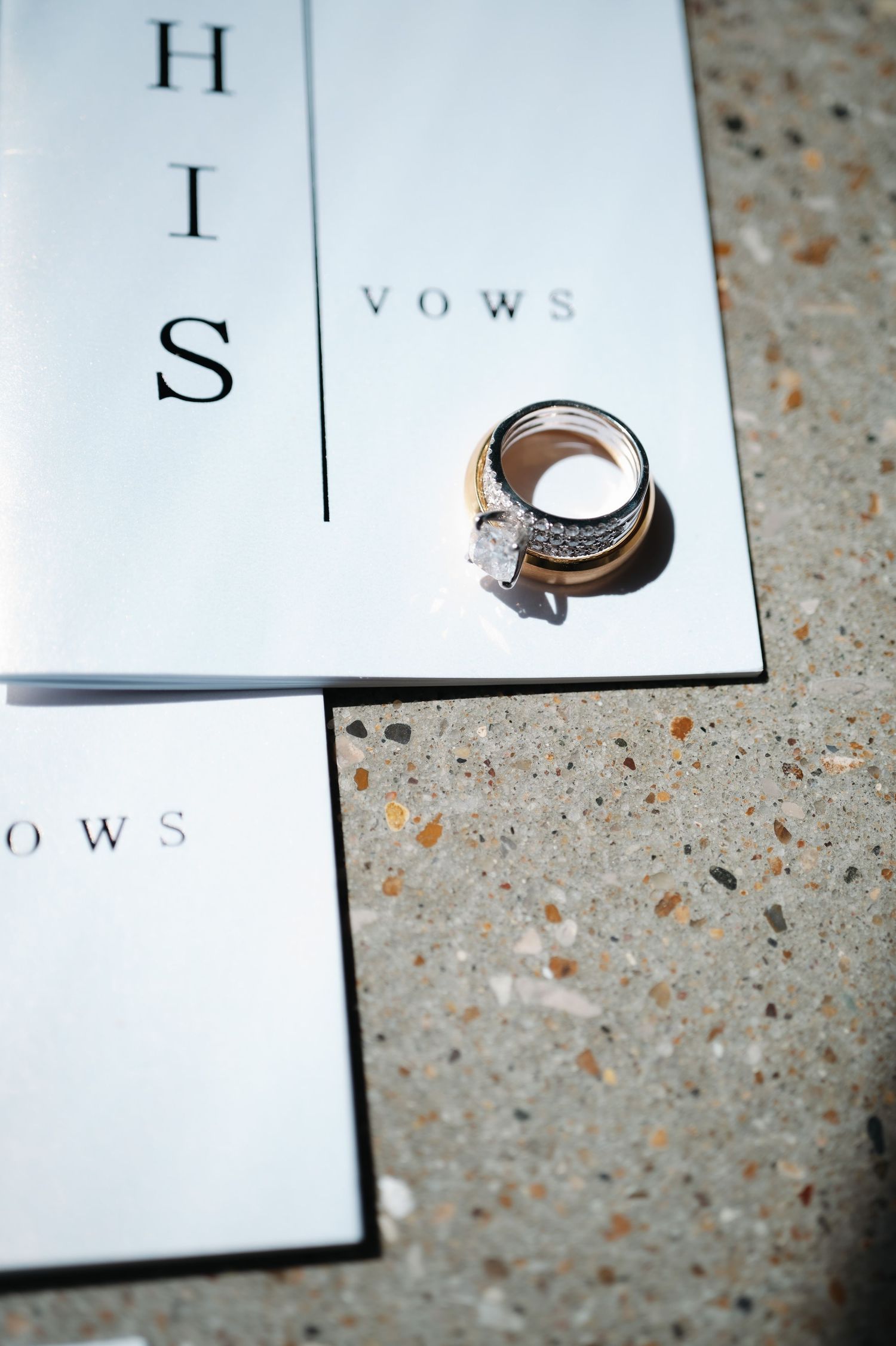Diamond engagement ring and wedding band resting on His and Hers vow booklets on stone surface.