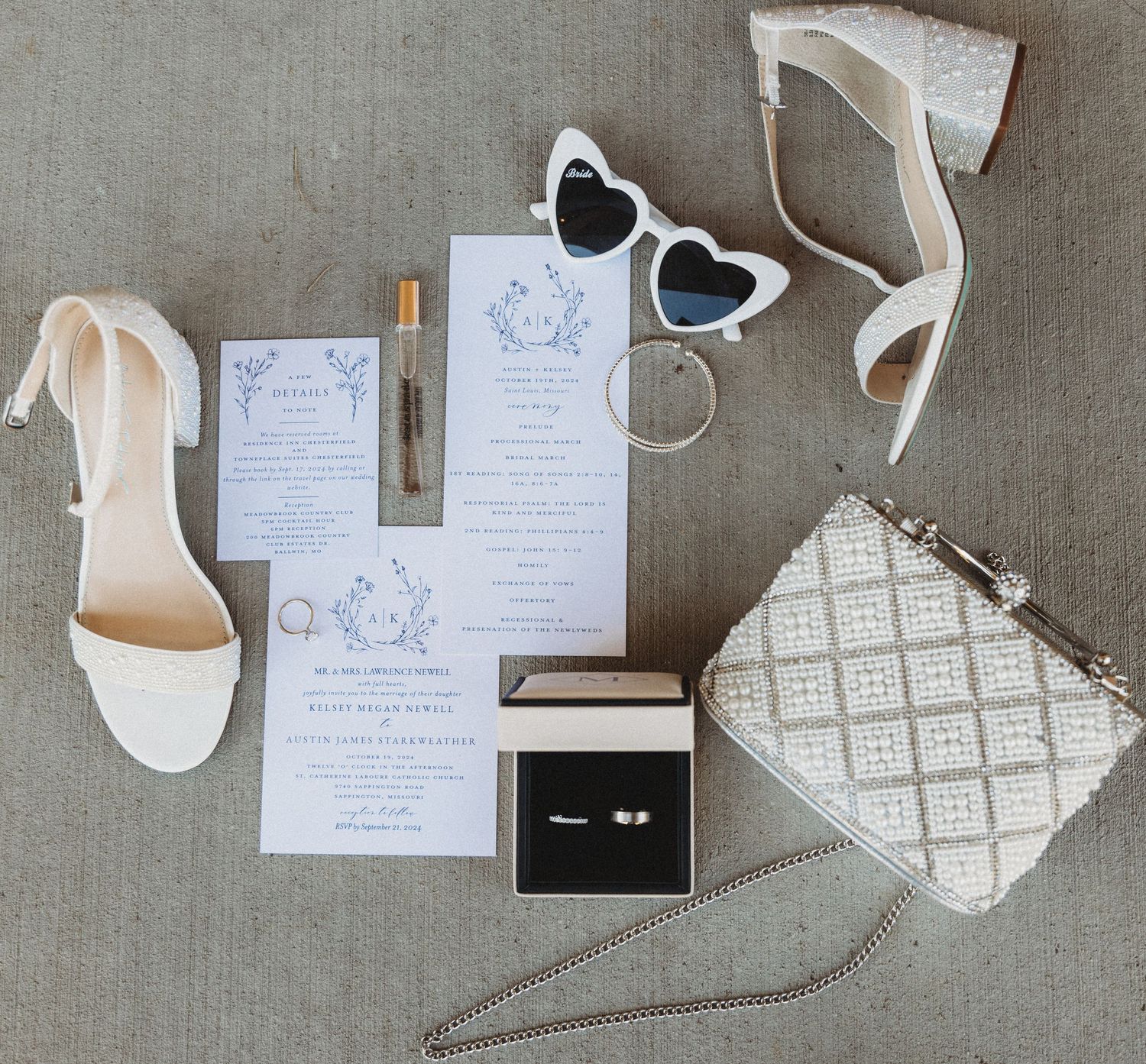 Flat lay of wedding stationery, white heels, heart sunglasses, perfume, ring box, and beaded clutch.