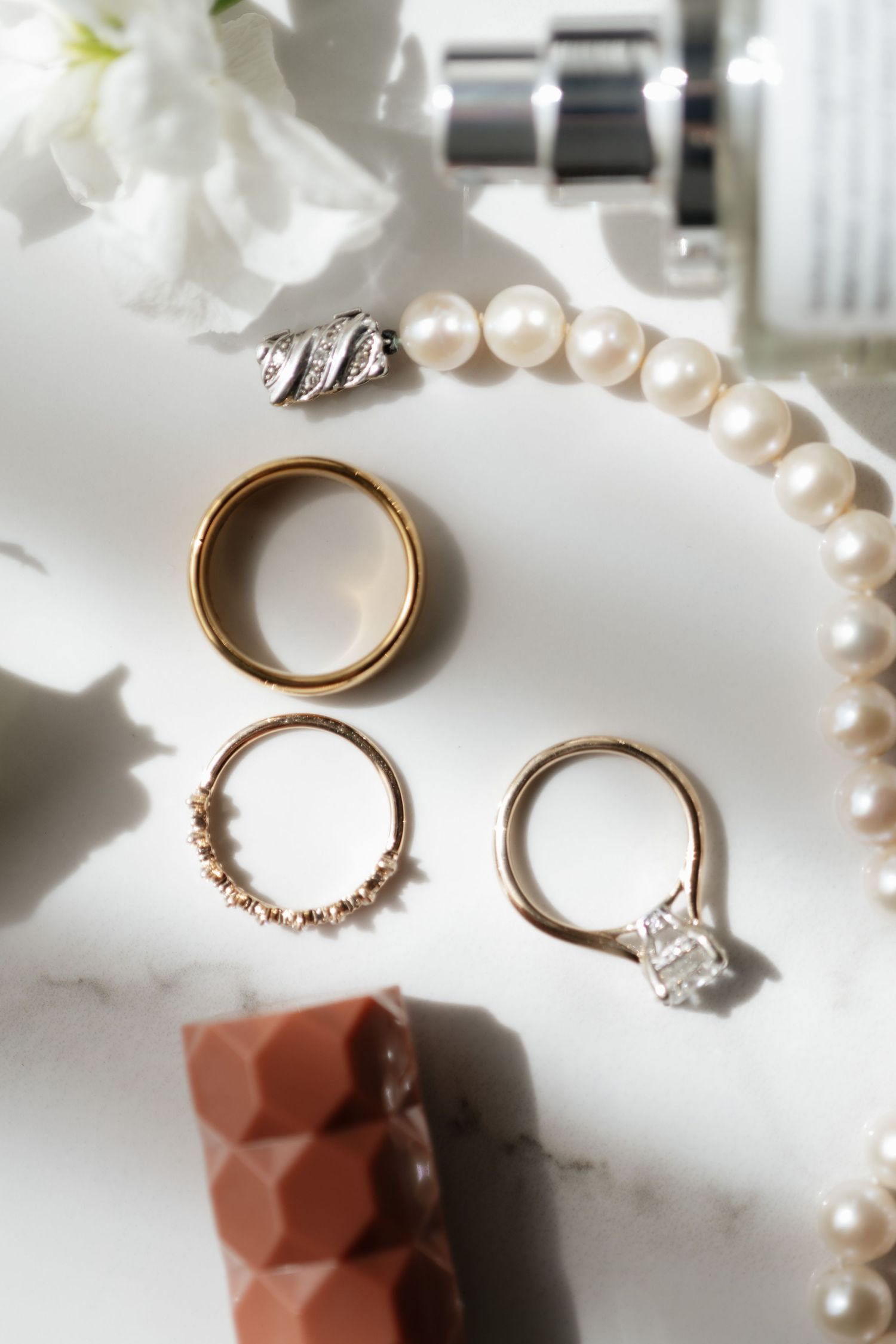 Flat lay of gold wedding bands, diamond engagement ring, pearl necklace, and earrings on marble.