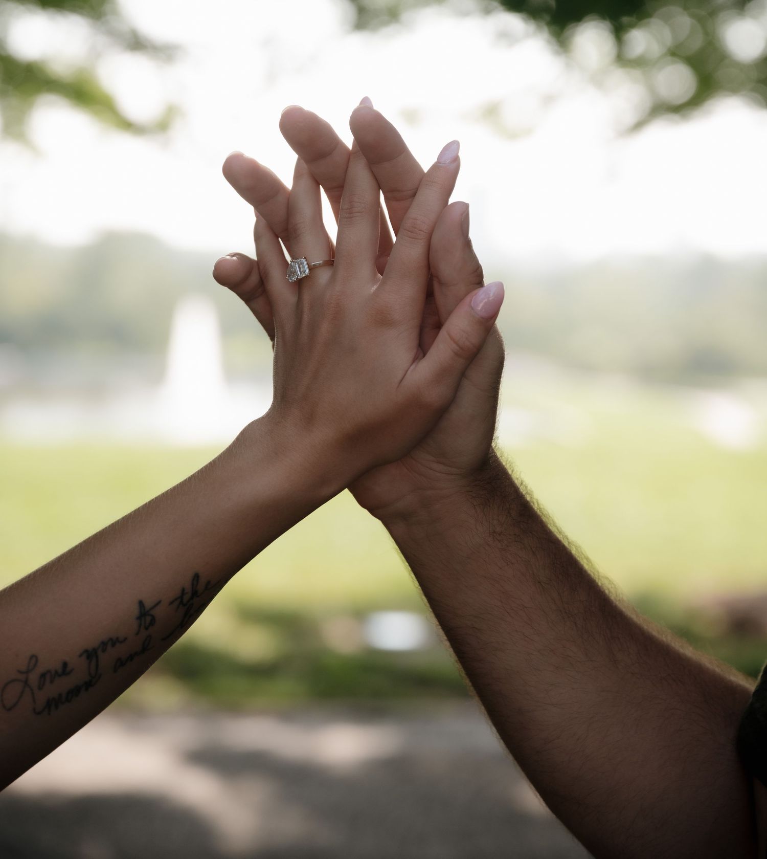 Couple holding hands outdoors, woman wearing diamond engagement ring with a cursive tattoo on her forearm.