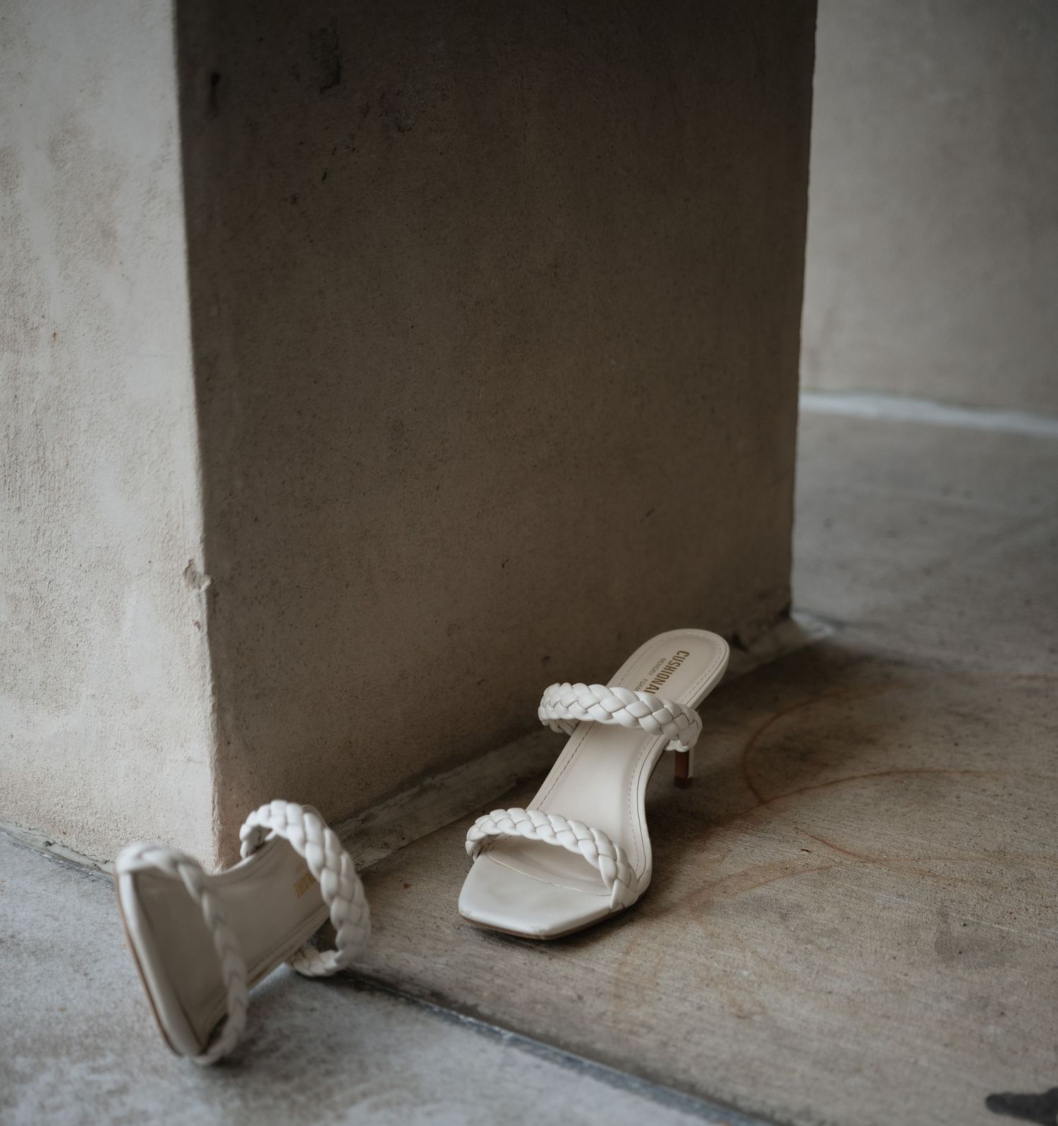 White braided strap bridal sandals resting against a concrete wall on stone floor.