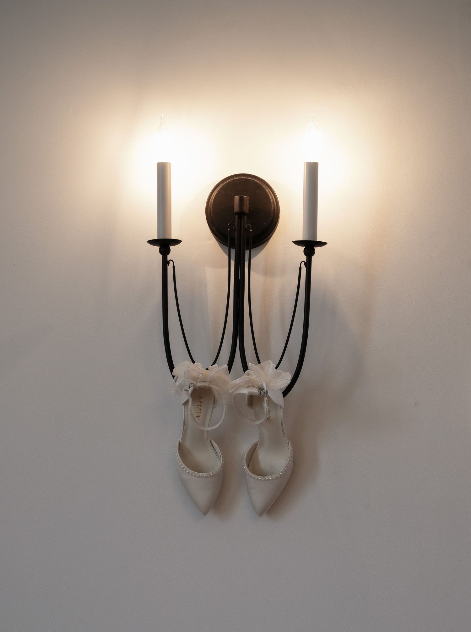 White pointed-toe bridal heels hanging from a two-candle iron wall sconce with glowing lights.