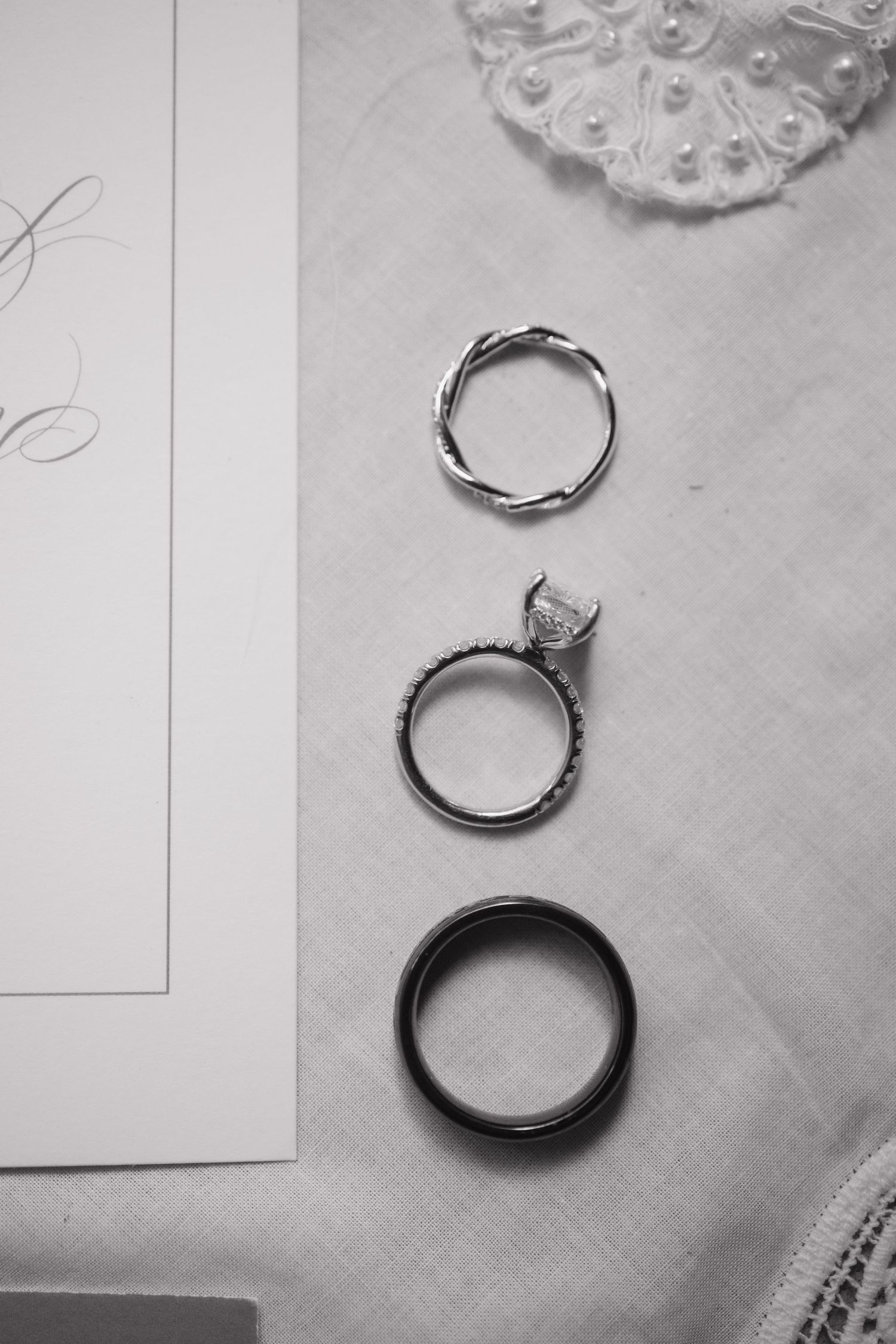 Black and white photo of three wedding rings arranged vertically beside calligraphy stationery and pearl bridal accessories.