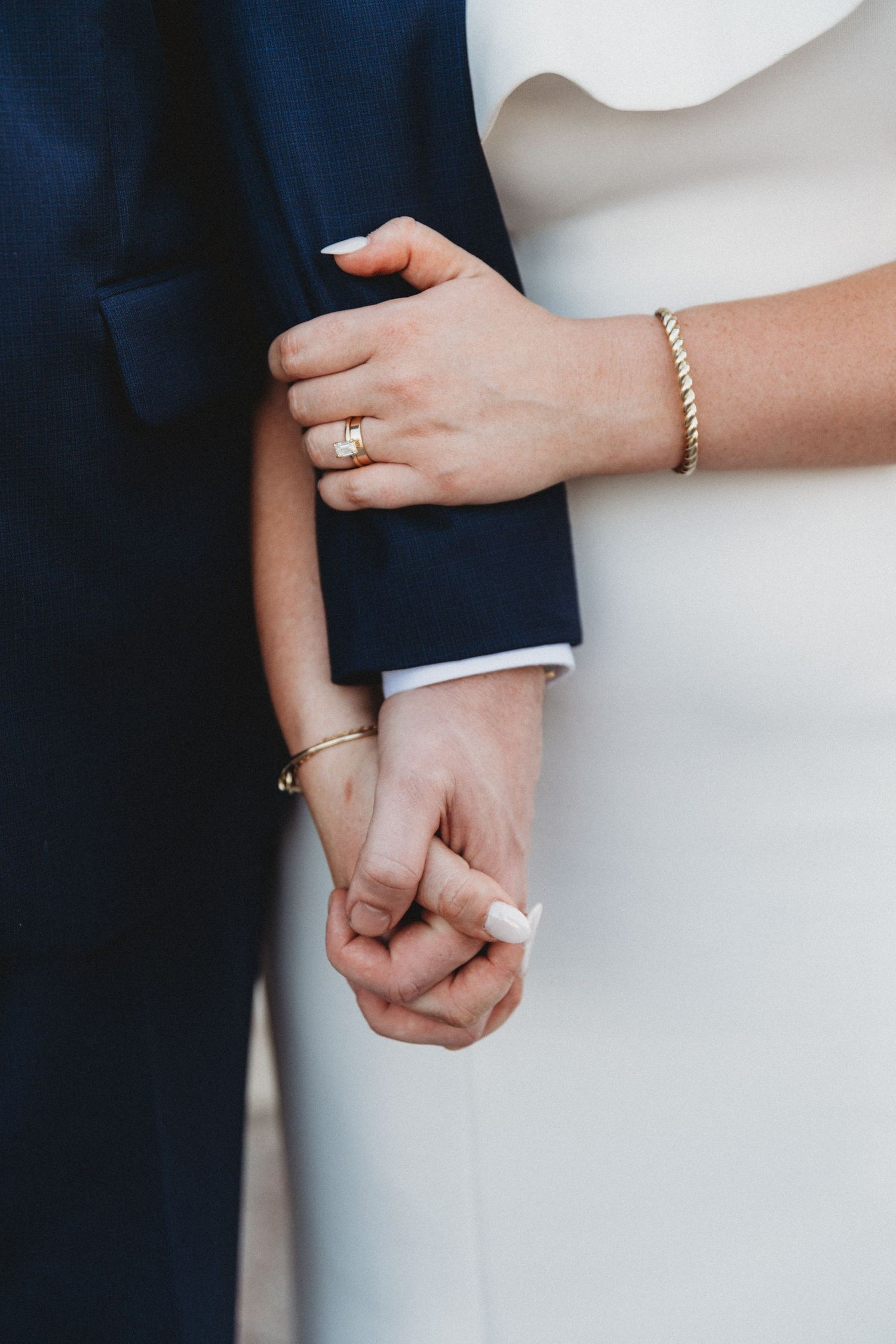 Close-up of bride and groom holding hands, bride wearing gold ring and bracelet in white dress.