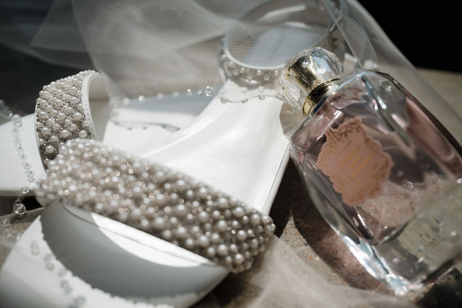 Close-up of pearl-embellished bridal sash, white veil, and pink perfume bottle on soft fabric.