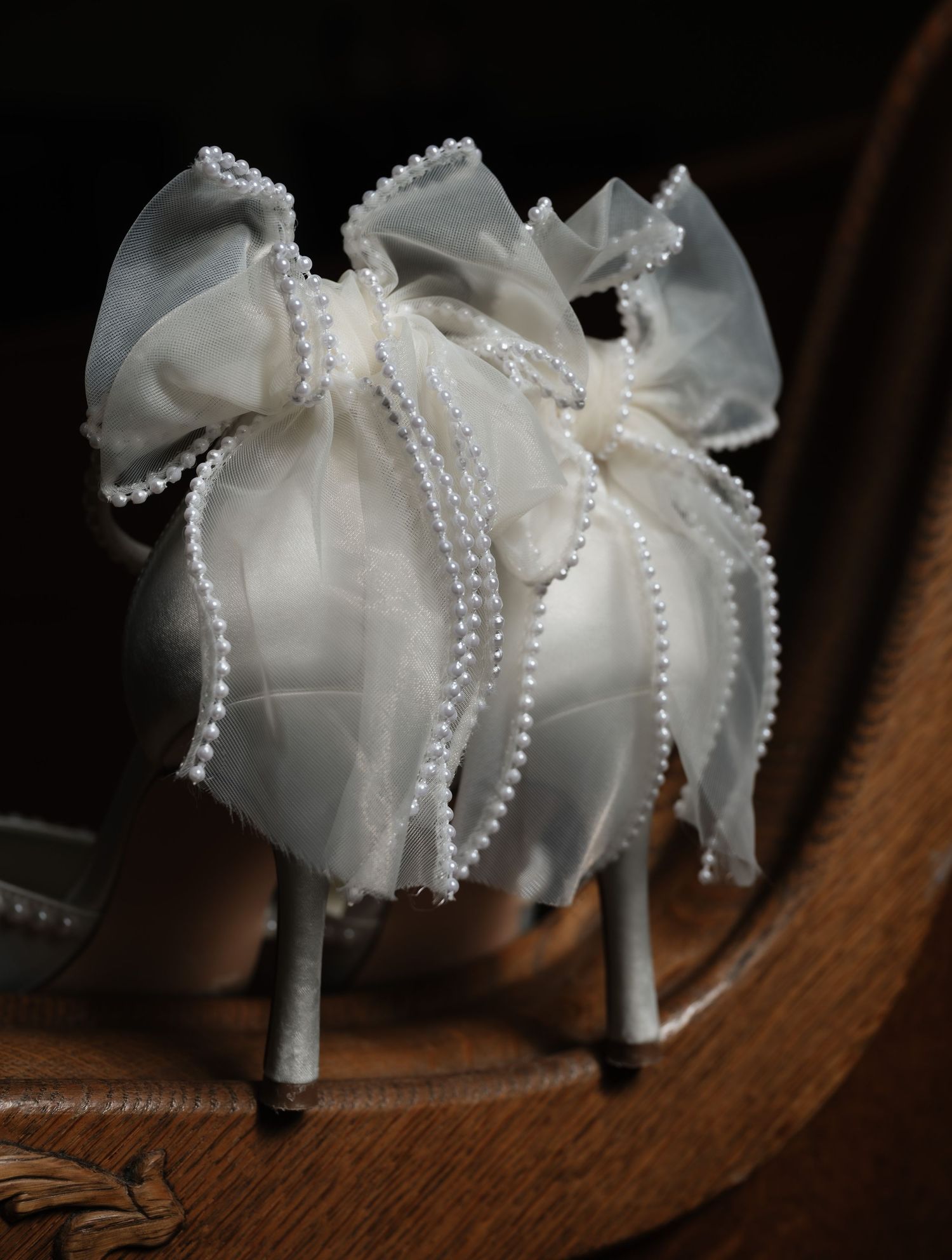 Elegant white bridal shoes adorned with delicate pearl embellishments and organza bows on a wooden surface.