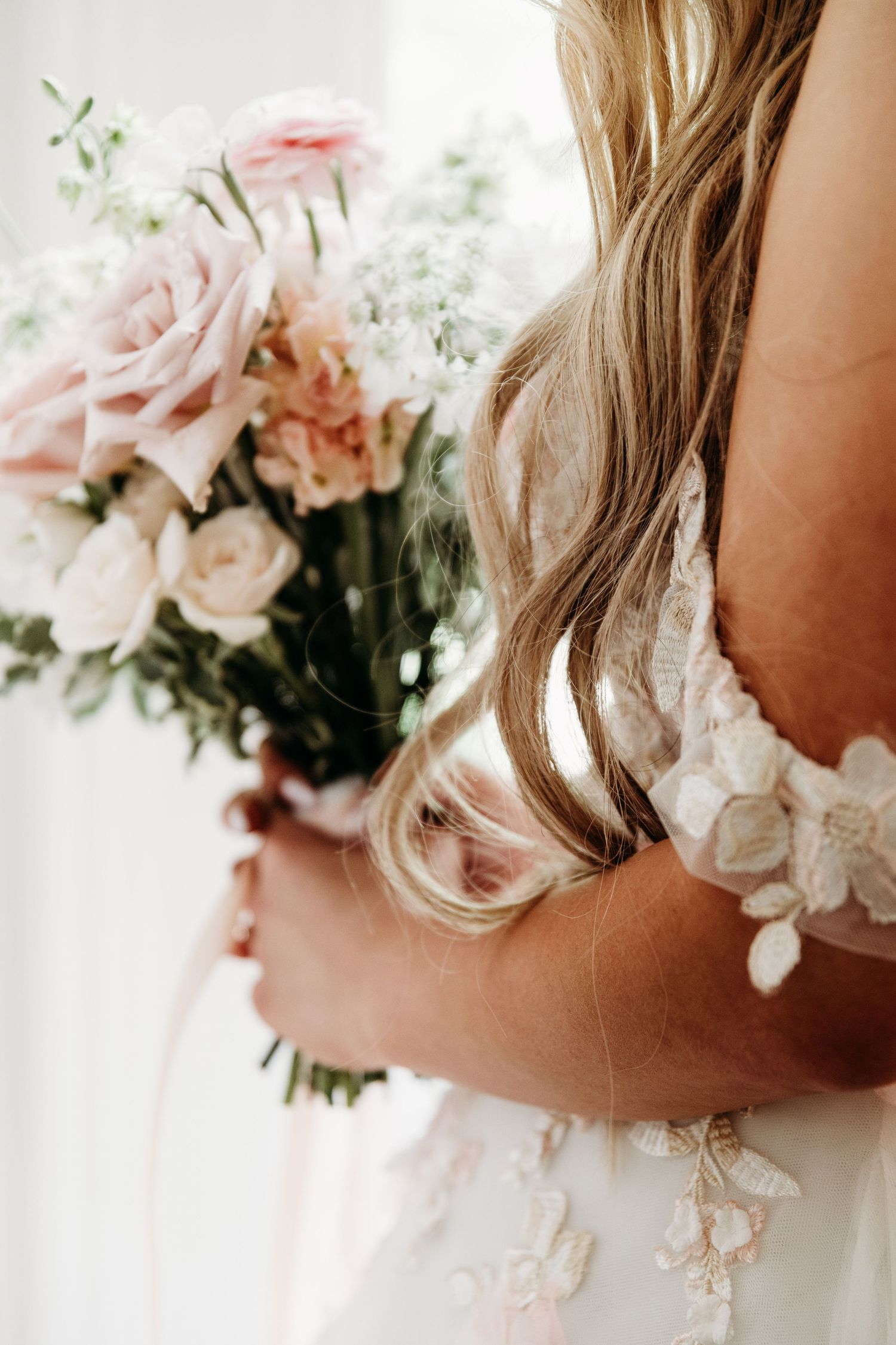 Blonde bride in floral lace wedding gown holding soft pink rose and carnation bouquet from behind.