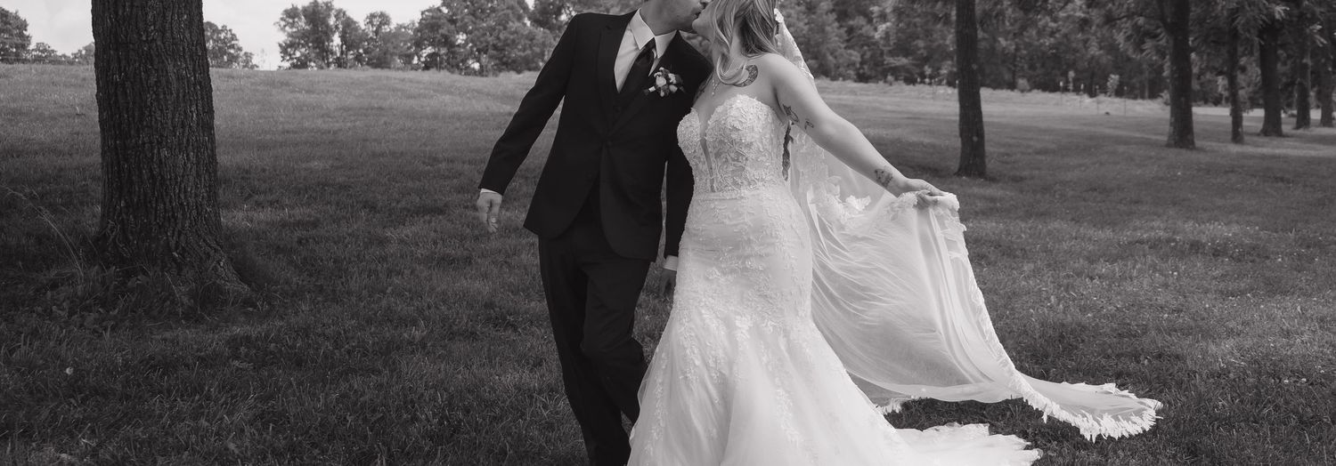 Black and white photo of a bride and groom walking hand in hand across a lush green field on their wedding day.