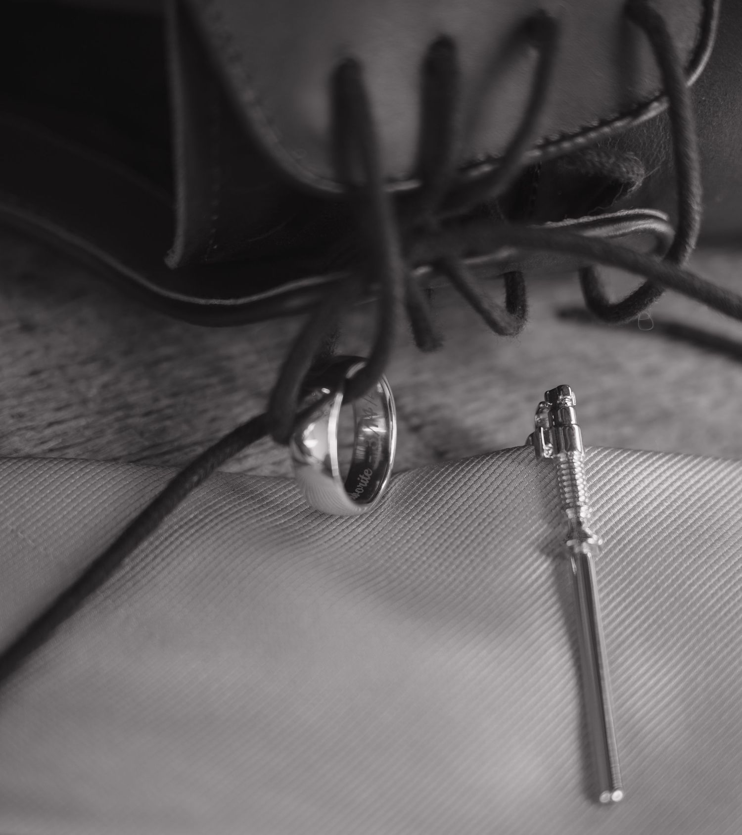 Black and white close-up of wedding ring and boutonniere pin beside leather dress shoe laces.