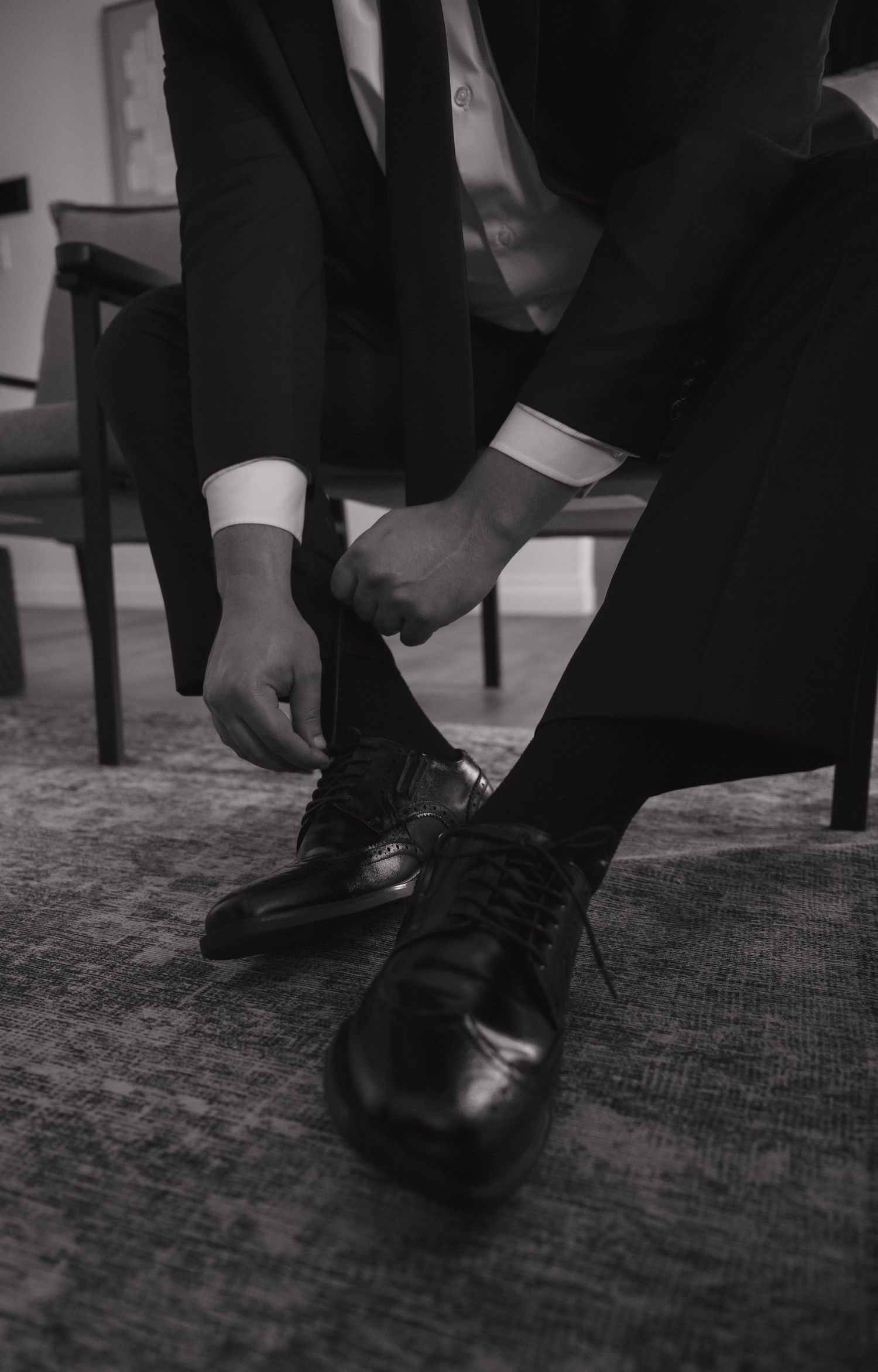 Groom in black suit tying dress shoes while seated, black and white wedding preparation photo.