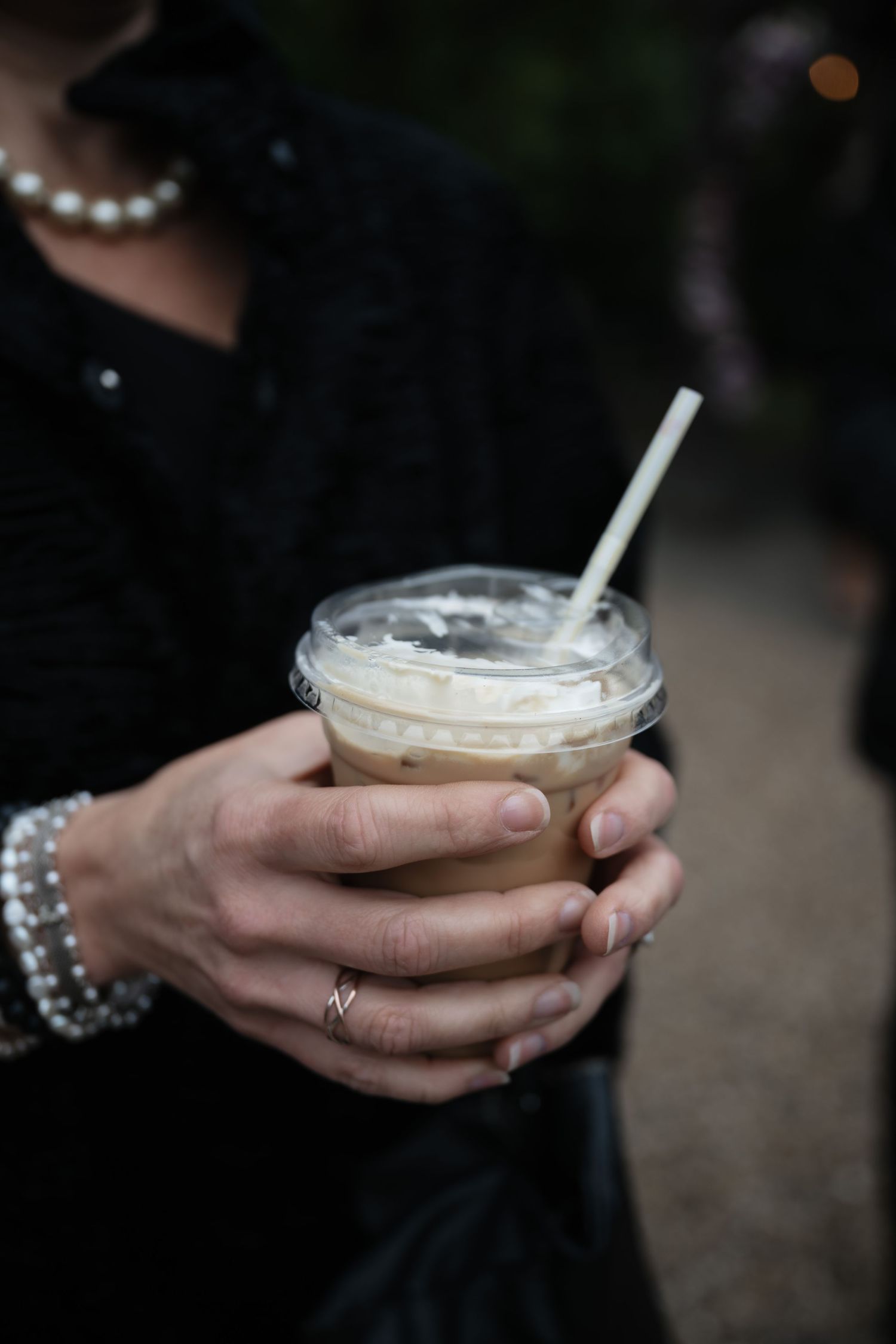 Hands wearing sparkly bracelets hold an iced coffee drink in a clear plastic cup with a white straw.