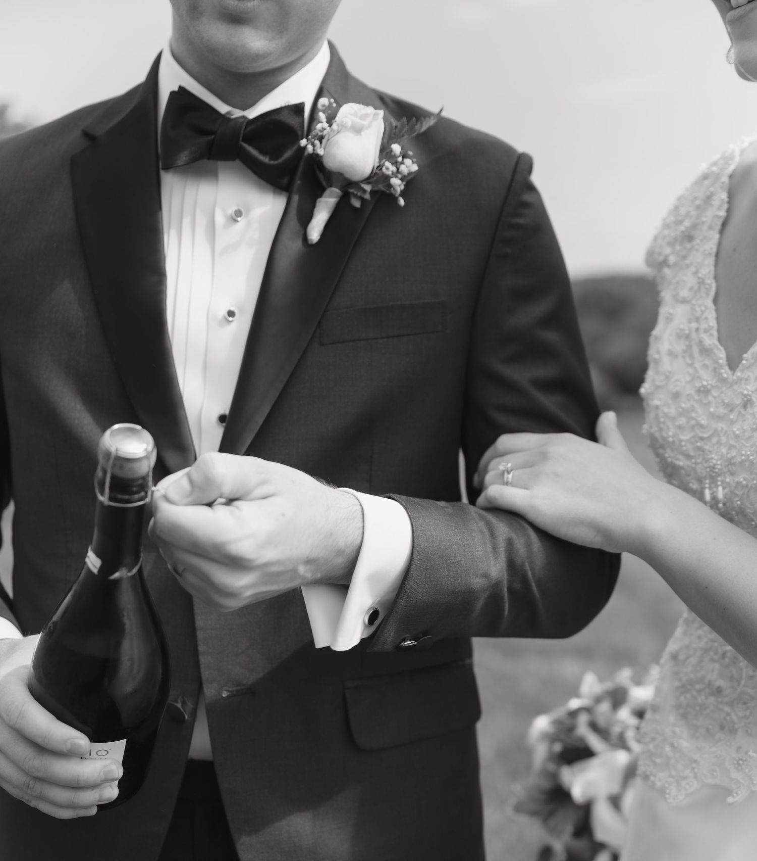 Black and white close-up of groom in tuxedo holding champagne bottle with bride in lace dress at wedding.
