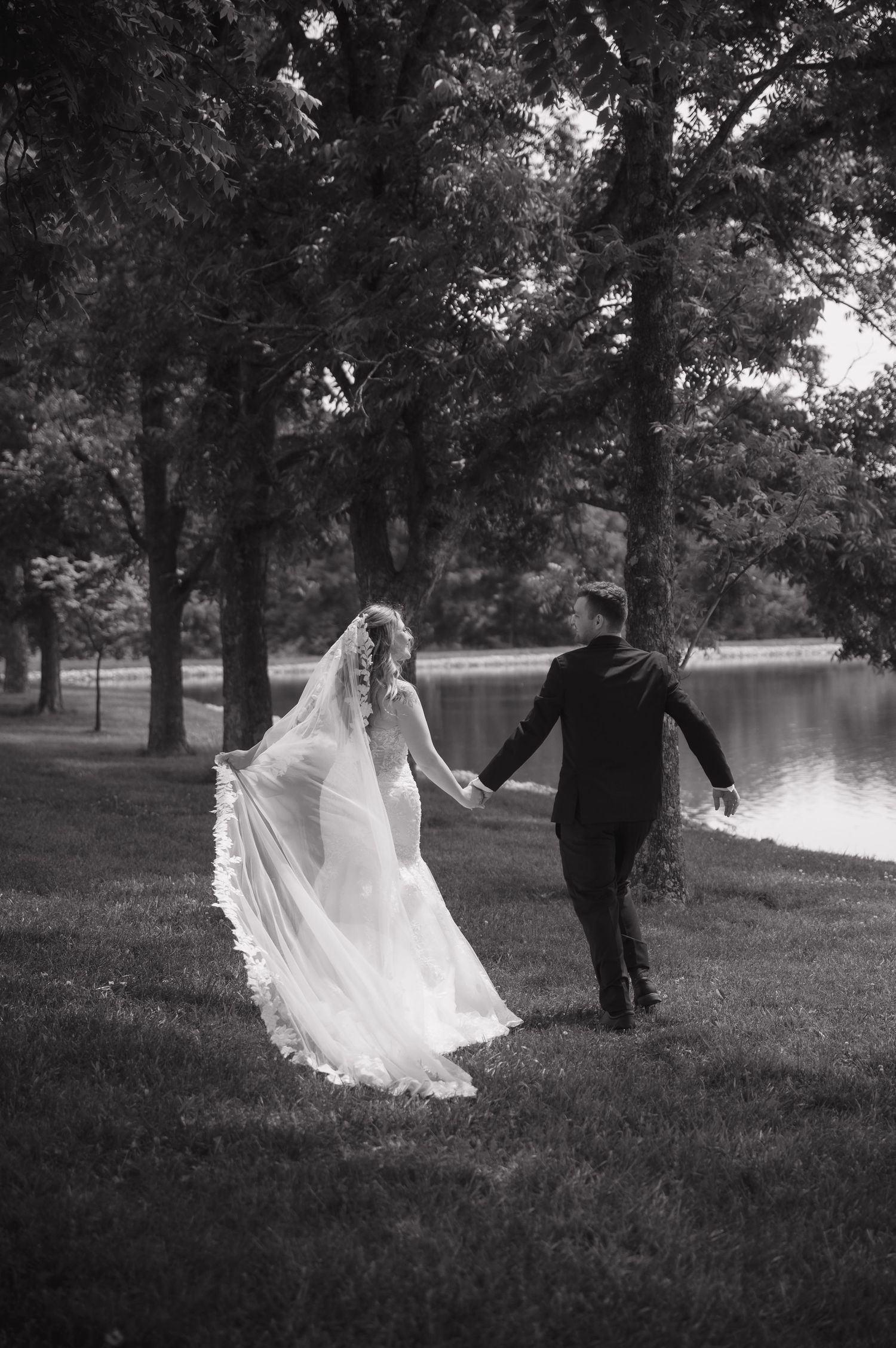 Black and white photo of bride and groom holding hands walking toward a lake surrounded by trees.
