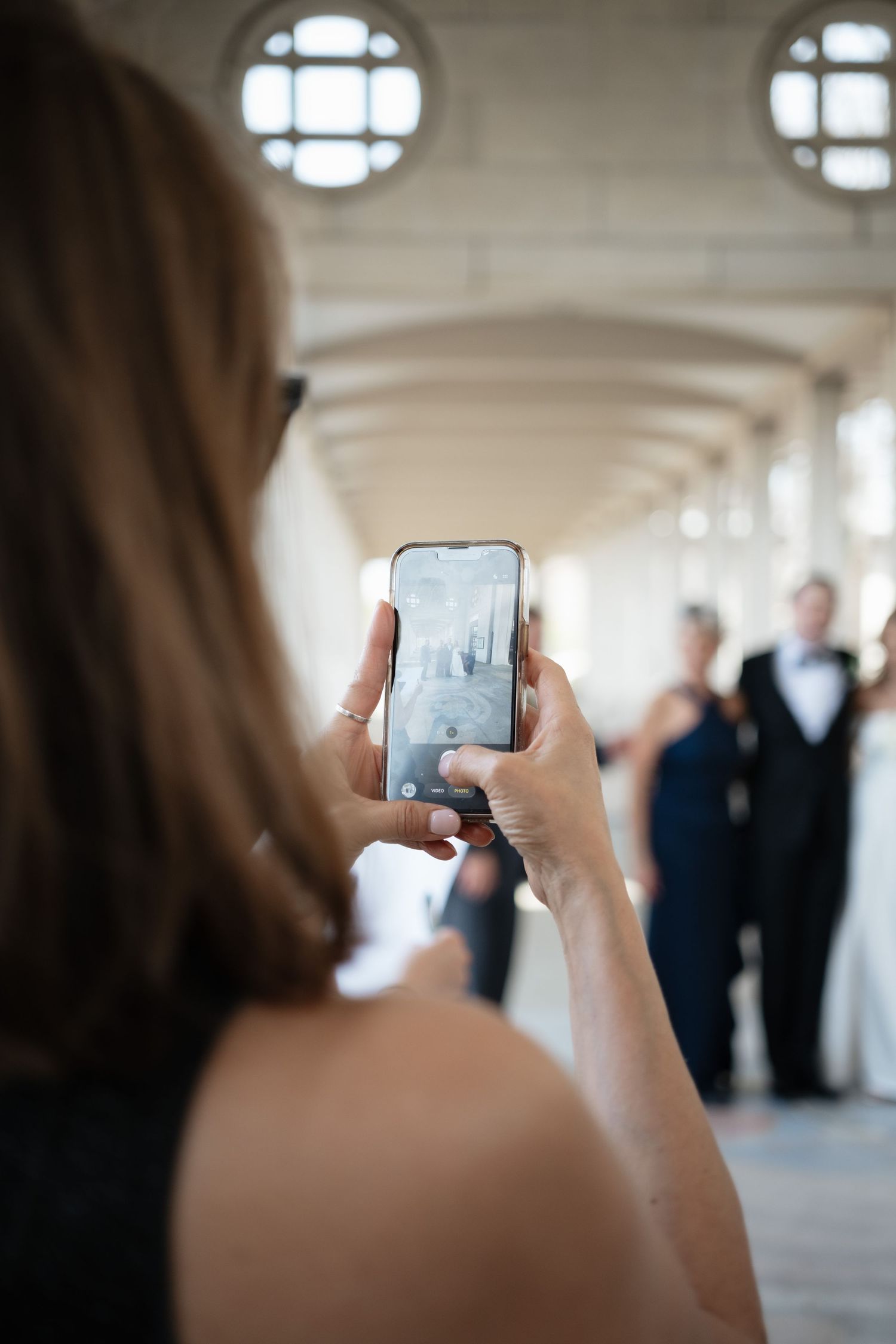 Woman photographing wedding guests with smartphone in elegant arched corridor.