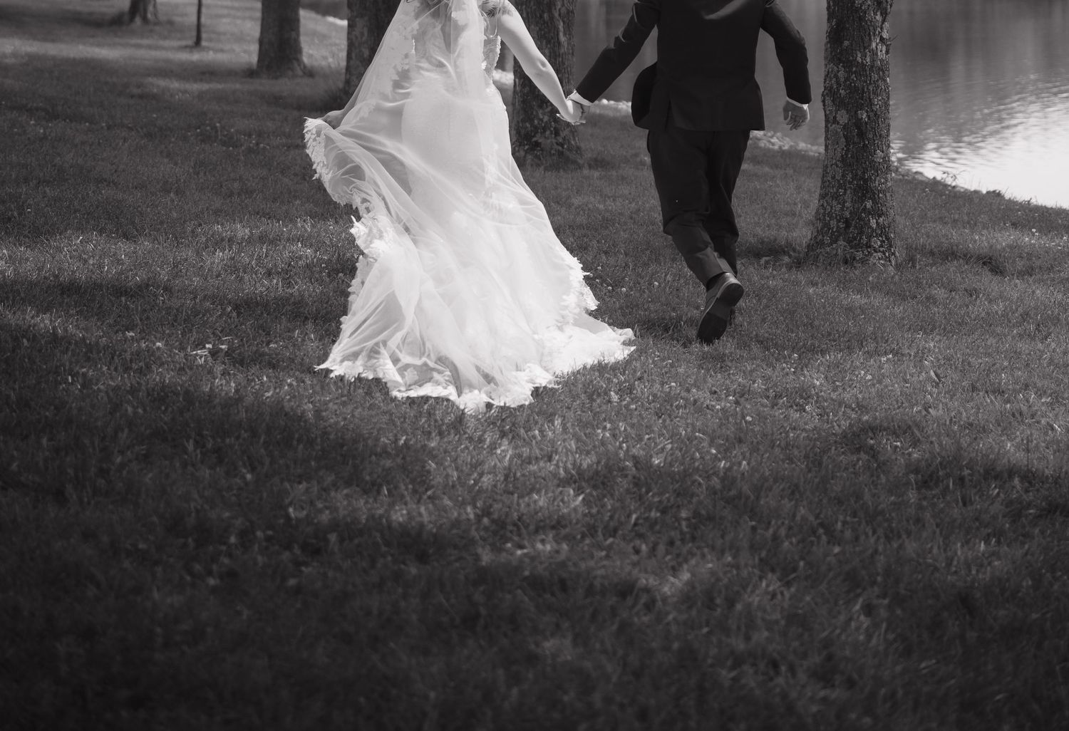 Black and white photo of bride and groom holding hands walking across green grass near a lake.