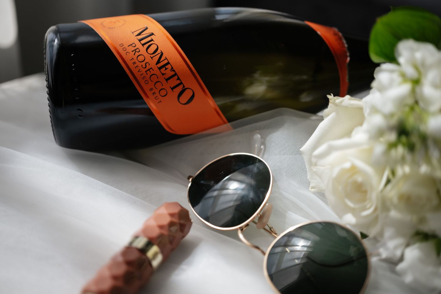 Monetto Prosecco bottle with aviator sunglasses and white roses on white fabric for wedding celebration.