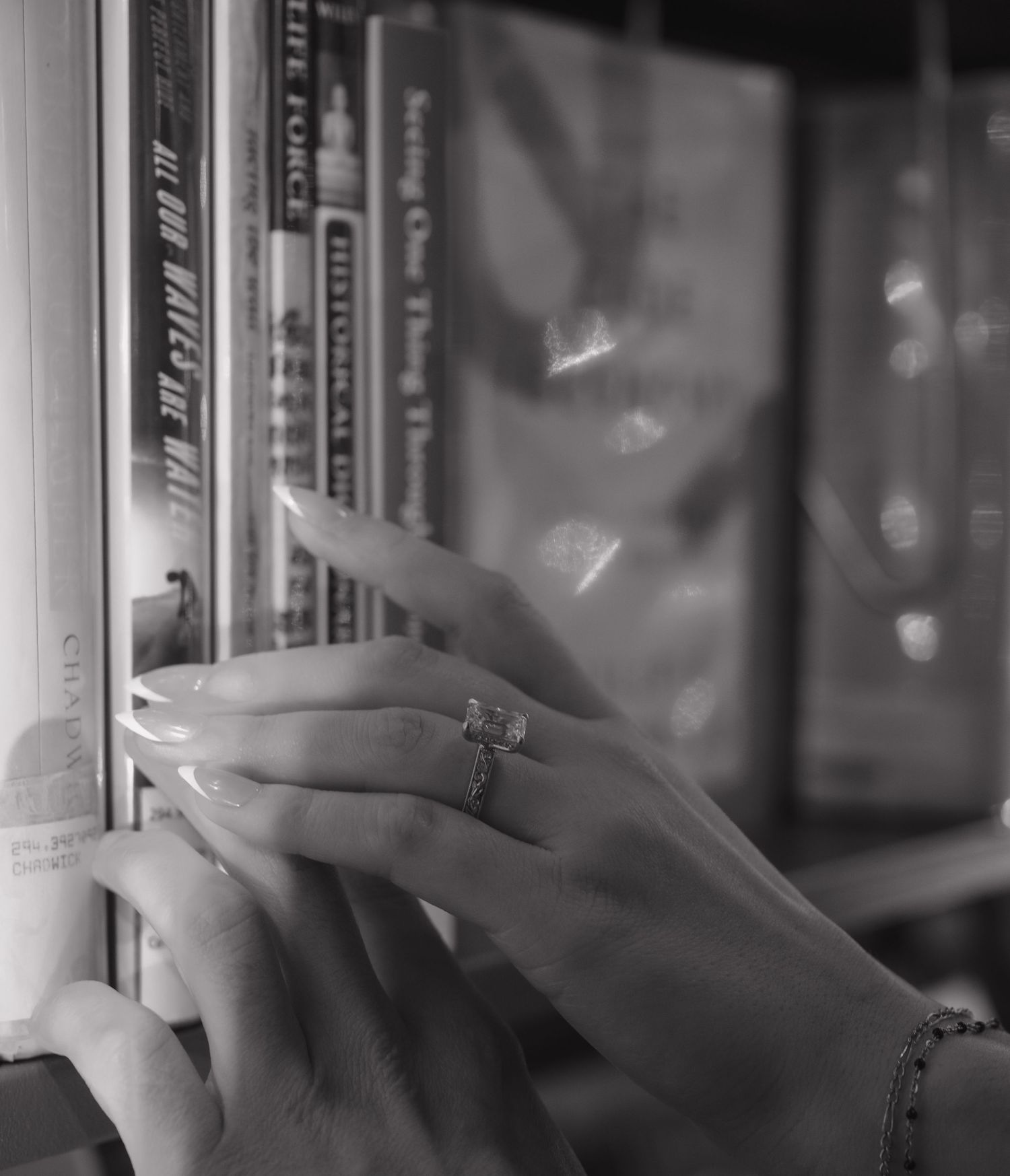 Close-up black and white photo of hand with diamond engagement ring browsing books on a shelf.