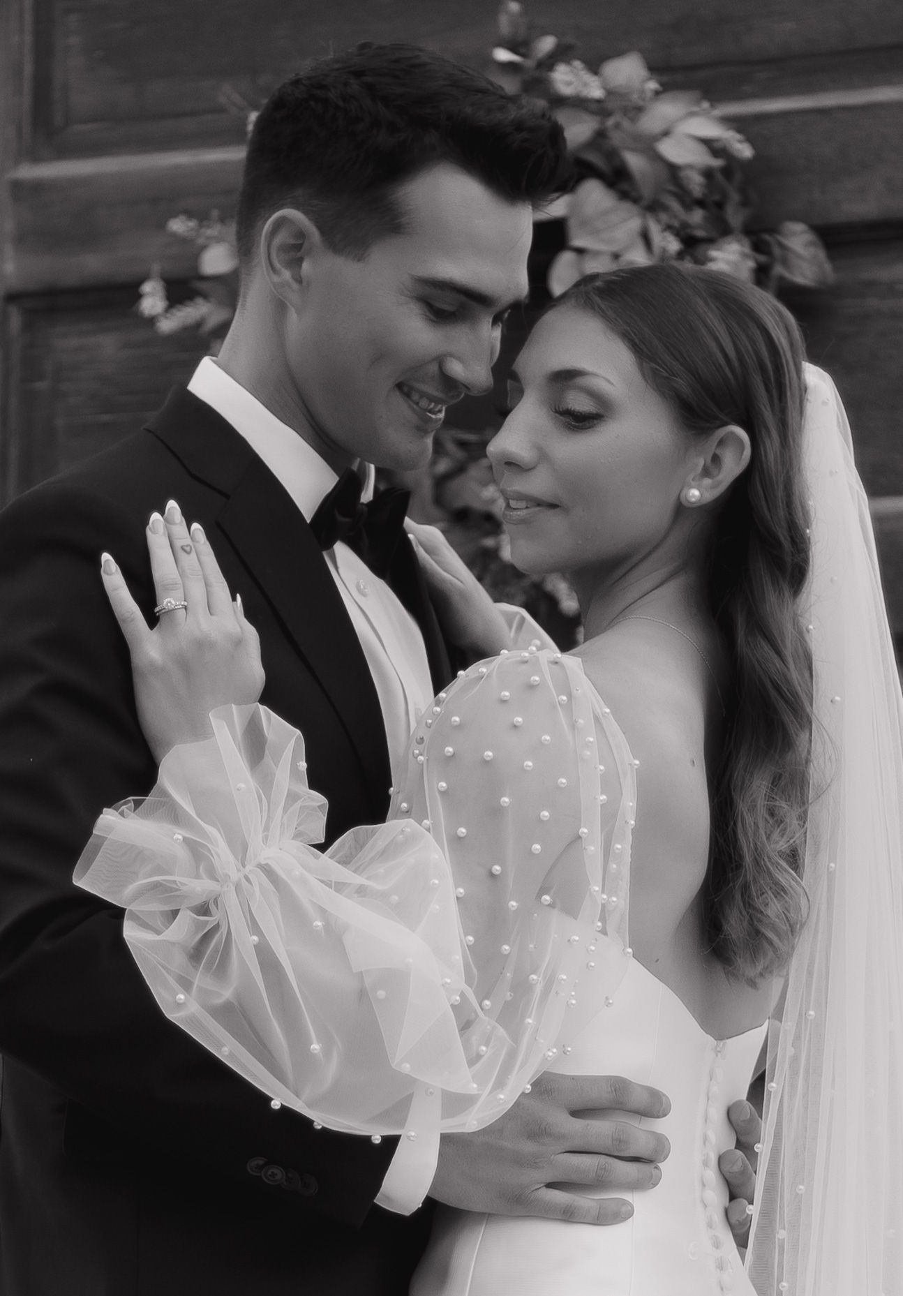 Black and white photo of bride and groom embracing at wedding ceremony, bride wearing pearl dress with bow sleeves.