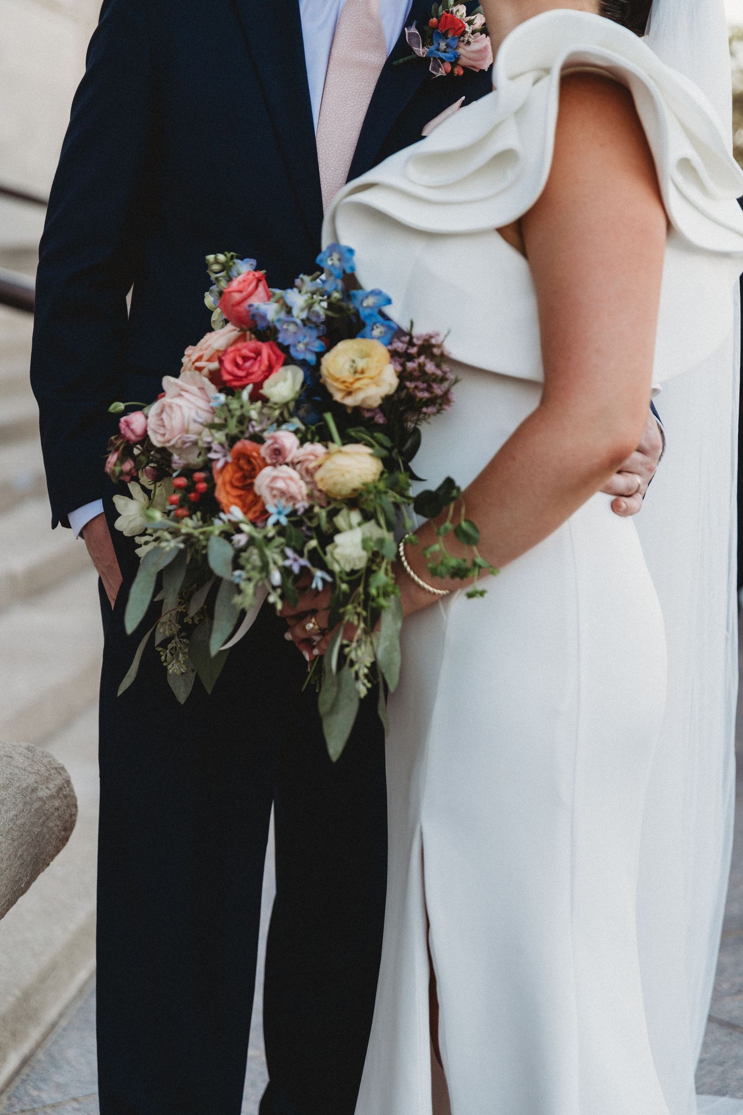 Bride in white sculptural off-shoulder dress holding vibrant garden bouquet with groom in navy suit.
